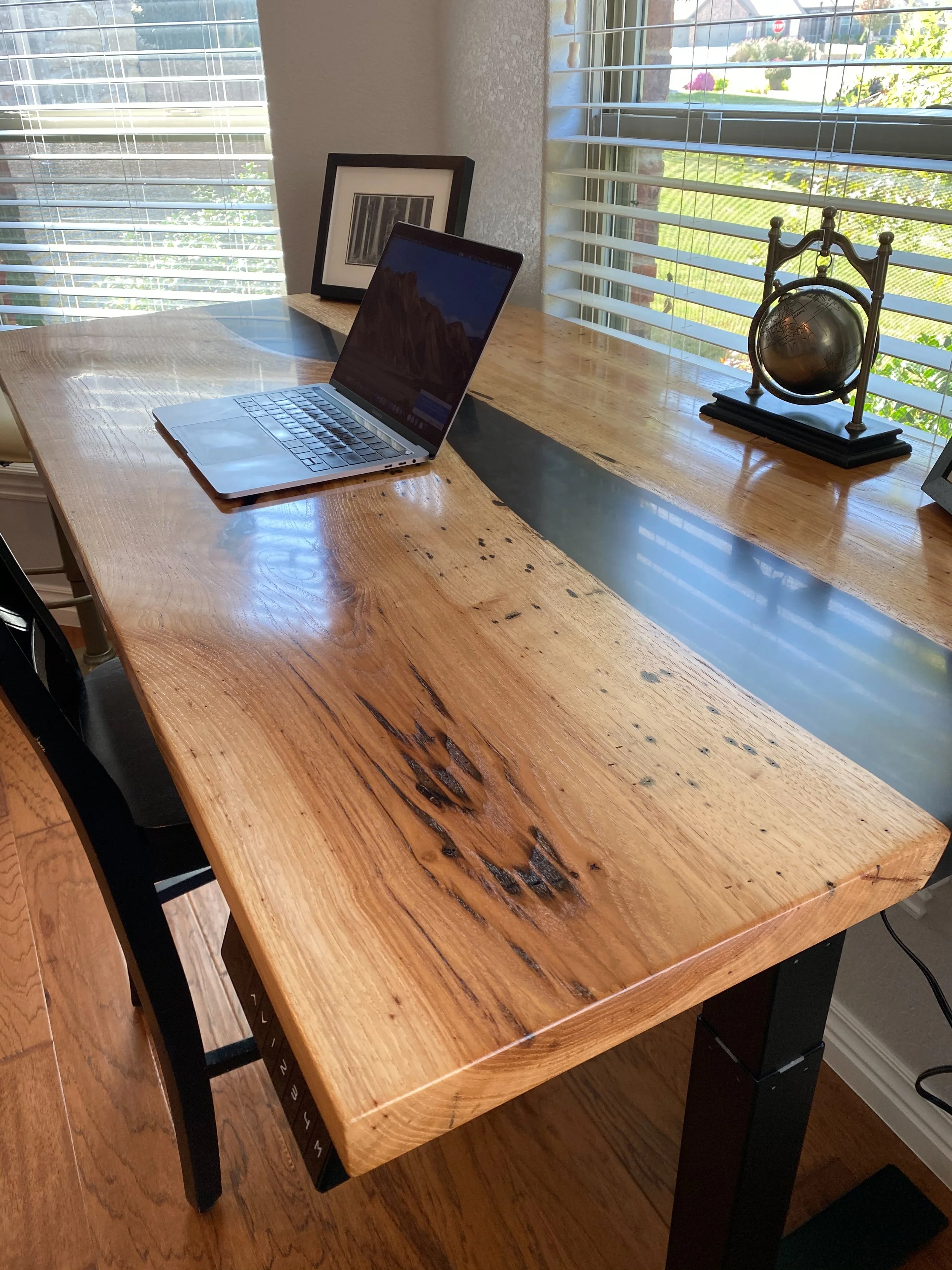 A Pecan and black onyx epoxy desk with a laptop, a framed photograph, and a decorative globe near a window with blinds, overlooking a sunny outdoor scene.