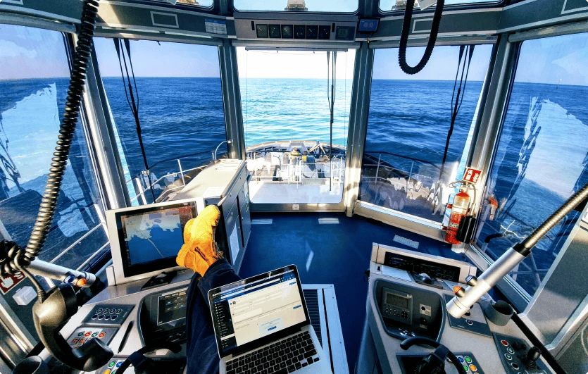 Interior of a ship's control room with large windows showing an ocean view, a desktop monitor, a laptop, a fire extinguisher, and a person with an orange glove operating the navigation equipment.