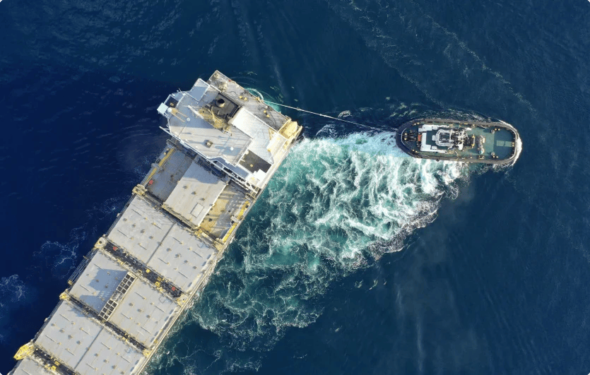 Overhead view of a large cargo ship being towed by a smaller tugboat in the ocean.
