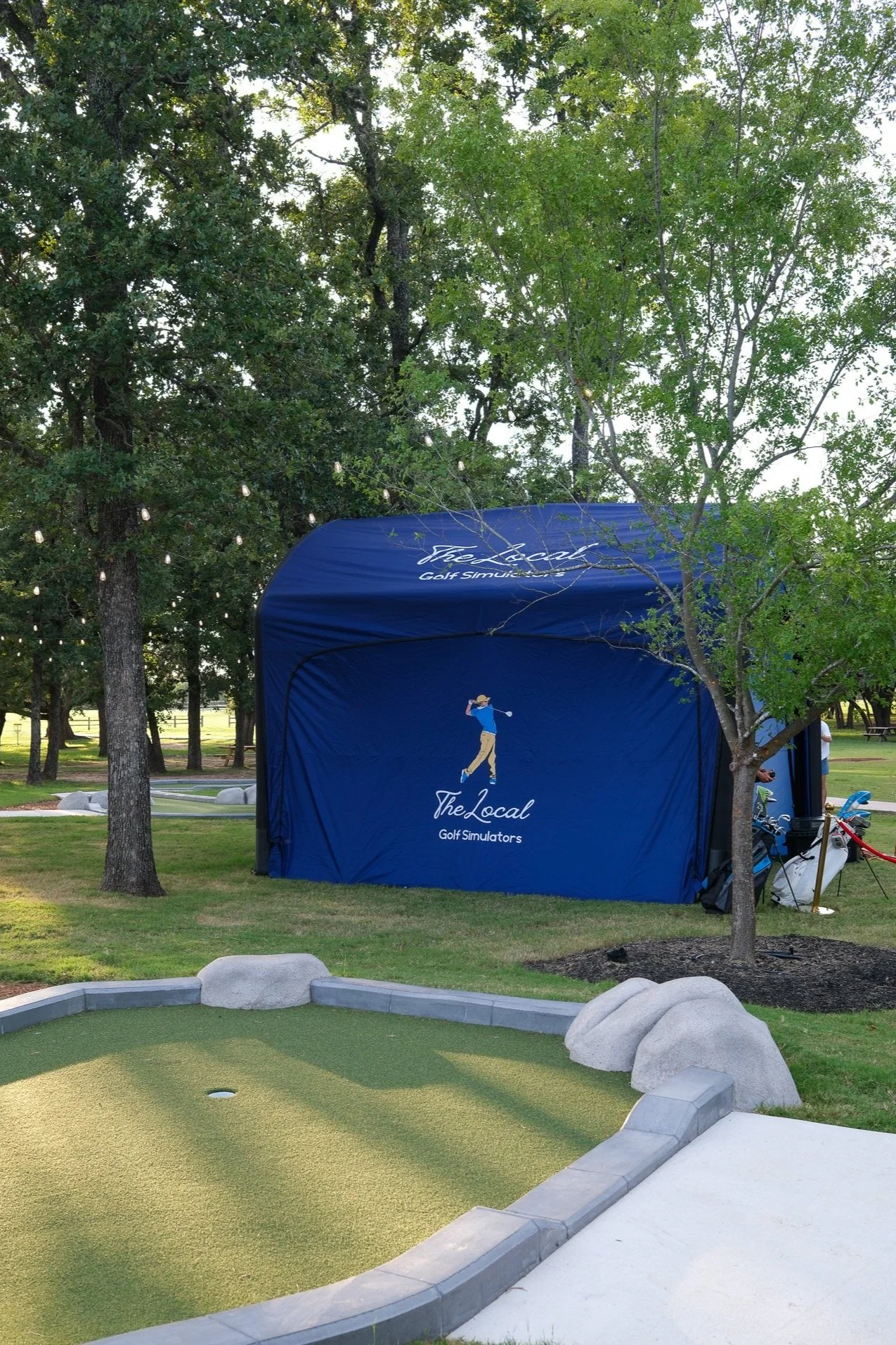 Outdoor mini golf course with a lush green putting area, trees, large rocks, and a blue tent with the logo and text for The Local Golf Simulators.