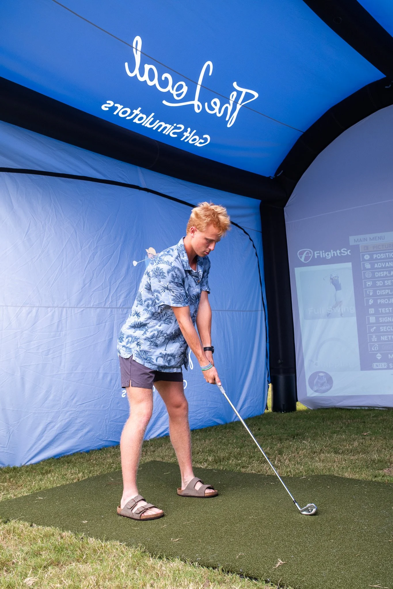 Young man with blonde hair wearing a blue Hawaiian shirt, shorts, and sandals practicing golf on an indoor simulation mat under a blue canopy with "Fairway" written on it.