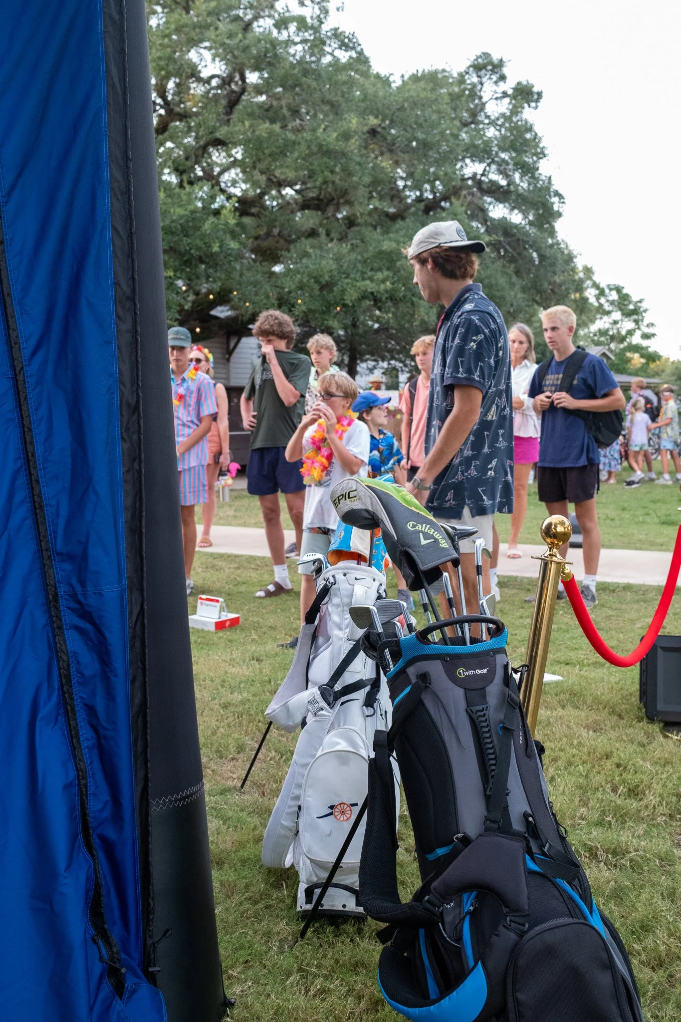 A group of young people, including children and teenagers, standing outside on a grassy area near a large blue object and a pair of golf bags filled with golf clubs, waiting or preparing for an event with trees in the background.