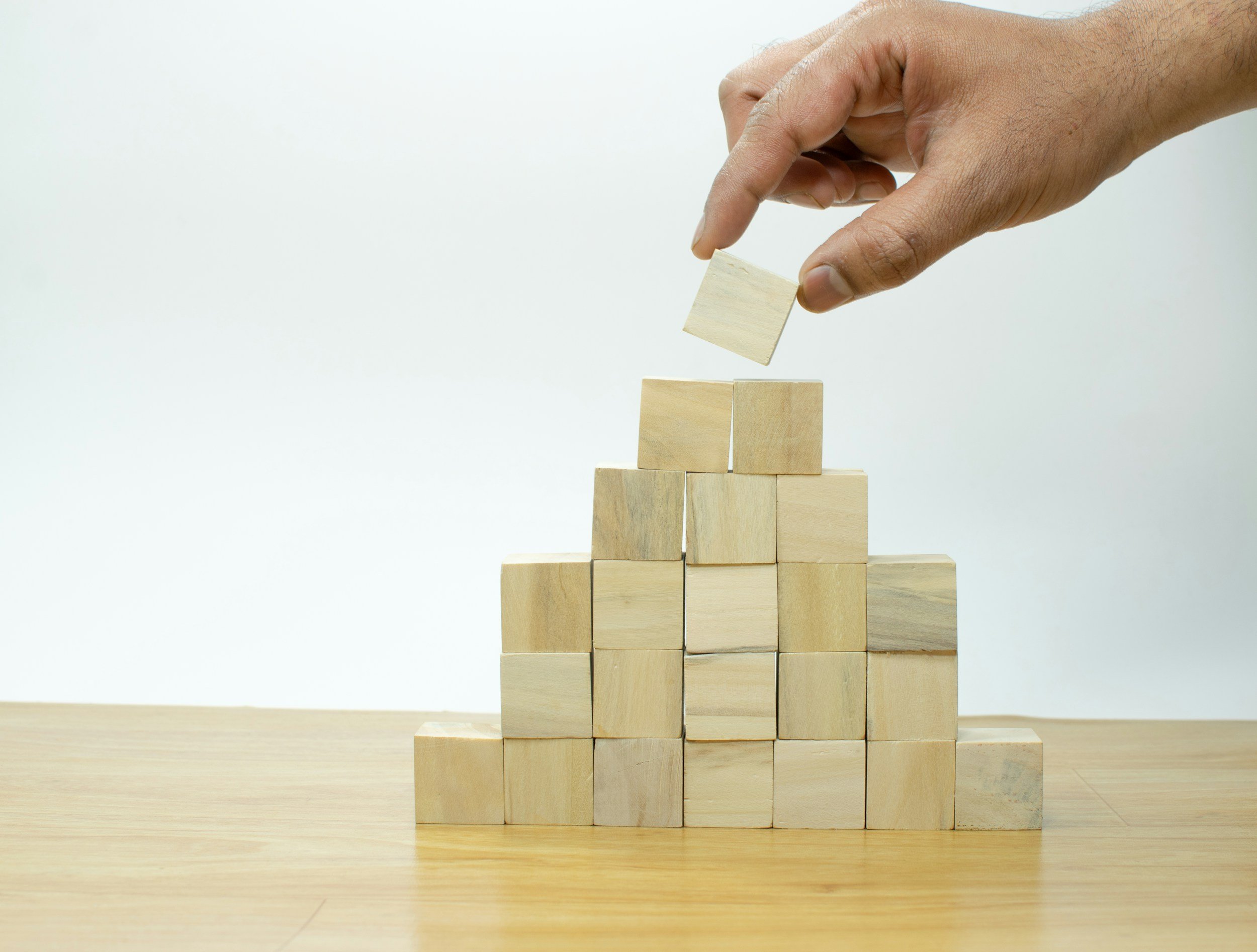 A hand placing a wooden block on top of a pyramid of wooden blocks on a wooden surface.