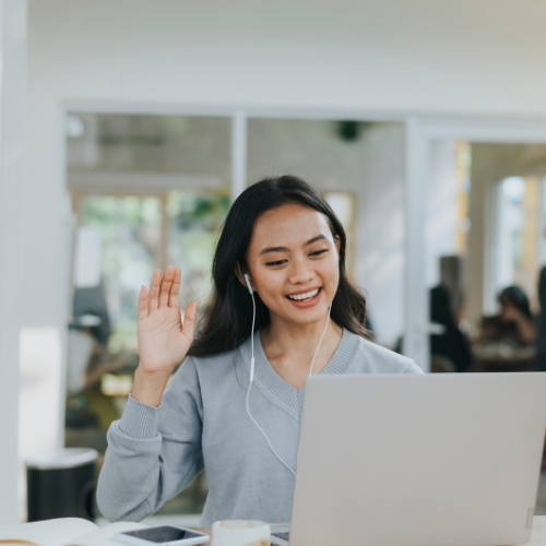Young woman with earphones smiling and waving at her laptop in a bright, modern room.