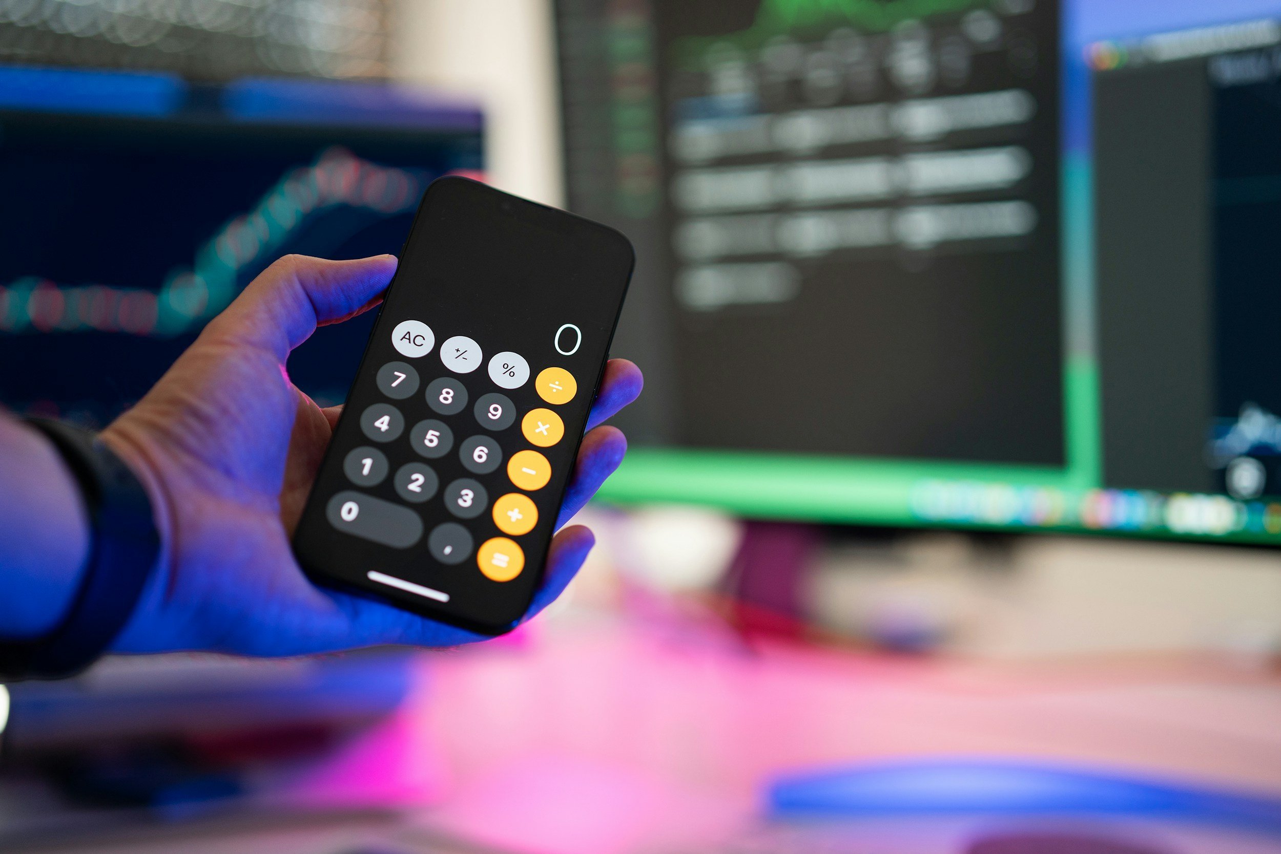 Person holding a smartphone with calculator app open, in front of a computer monitor displaying stock market data.