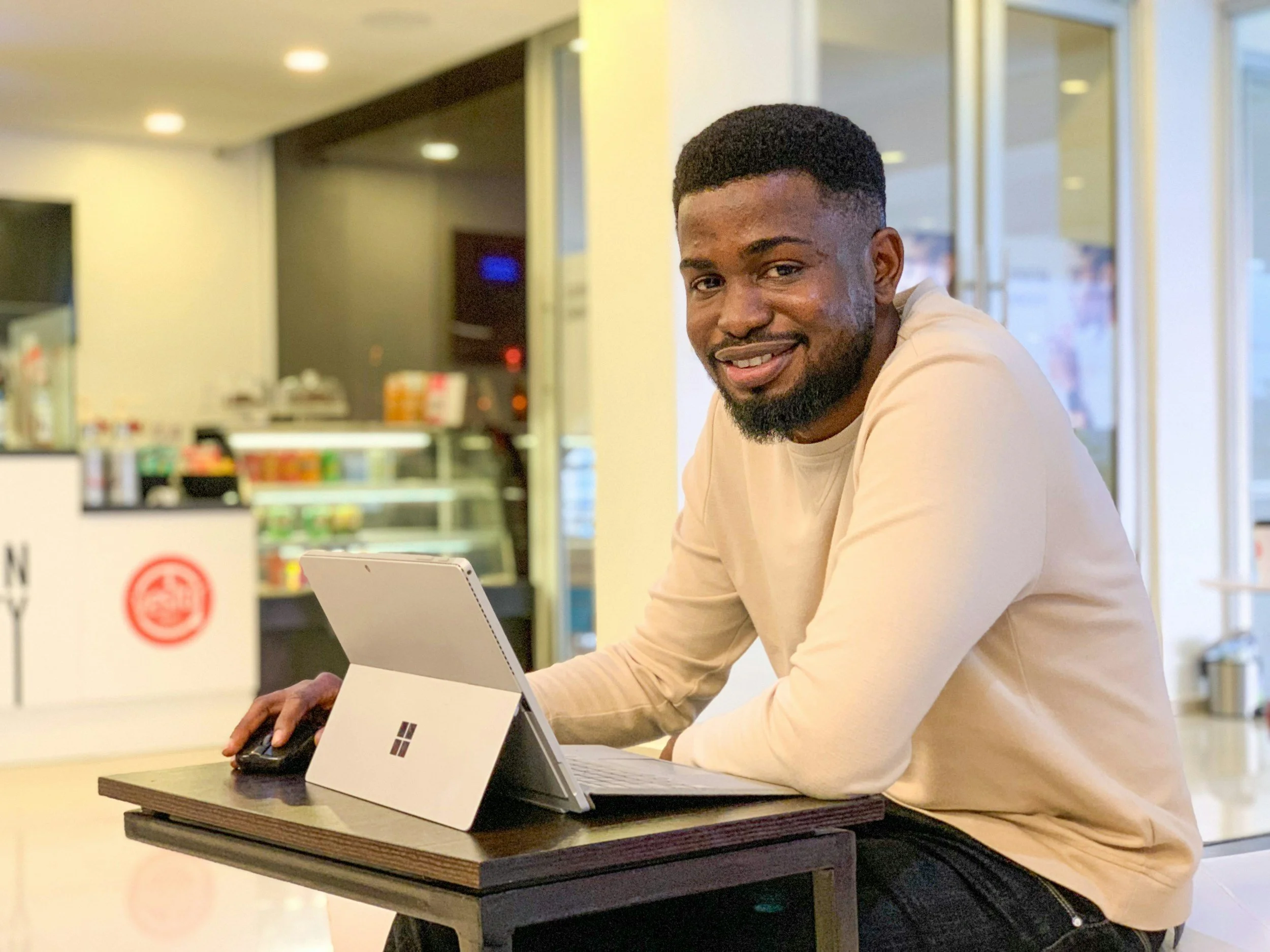 A smiling man sitting at a table with a Microsoft Surface tablet in a bright indoor space, possibly a café or coffee shop.
