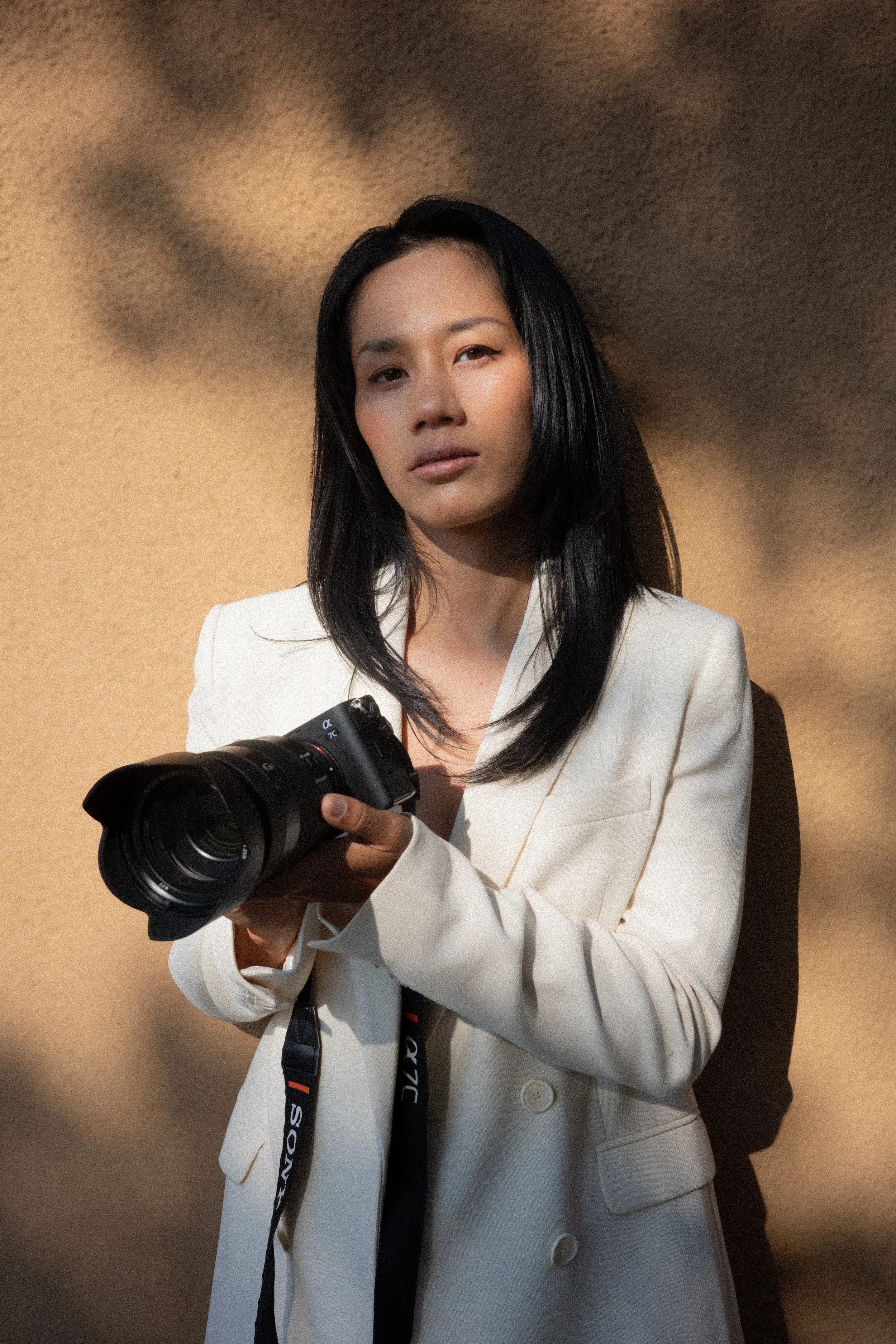 A woman wearing a white blazer holding a professional camera, with shadows on a beige background.