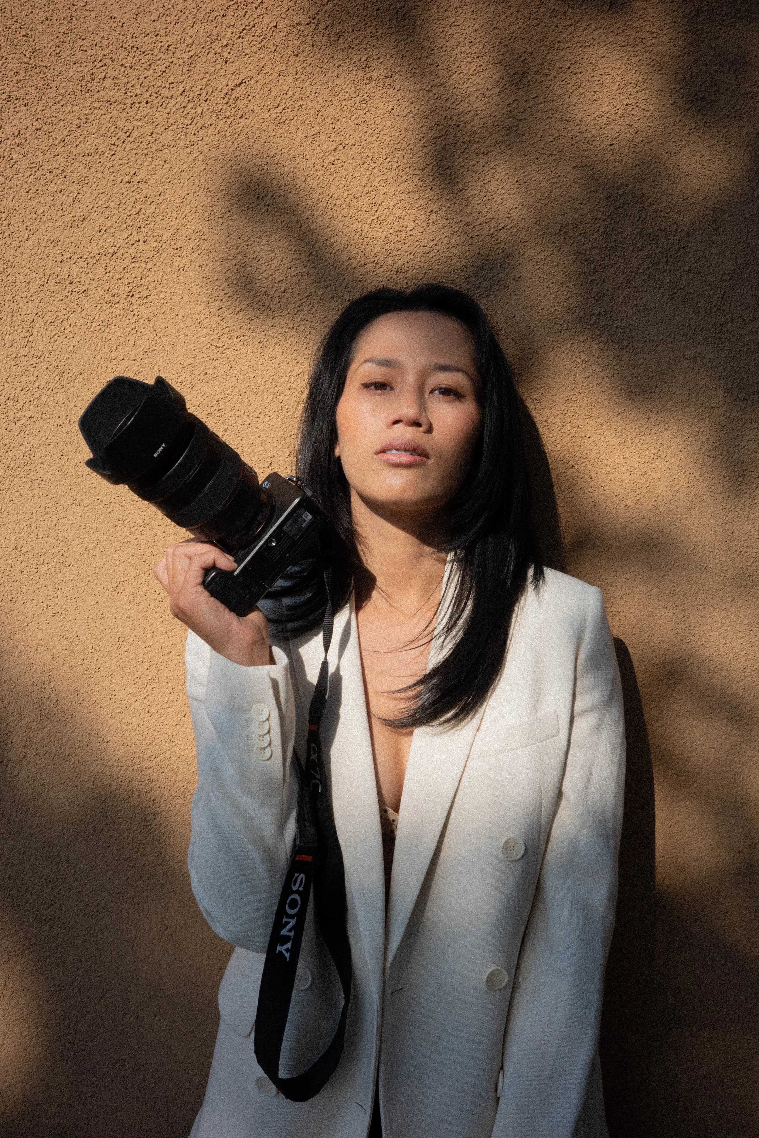 A woman with black hair wearing a white blazer holds a camera over her shoulder against a textured beige wall with shadows.