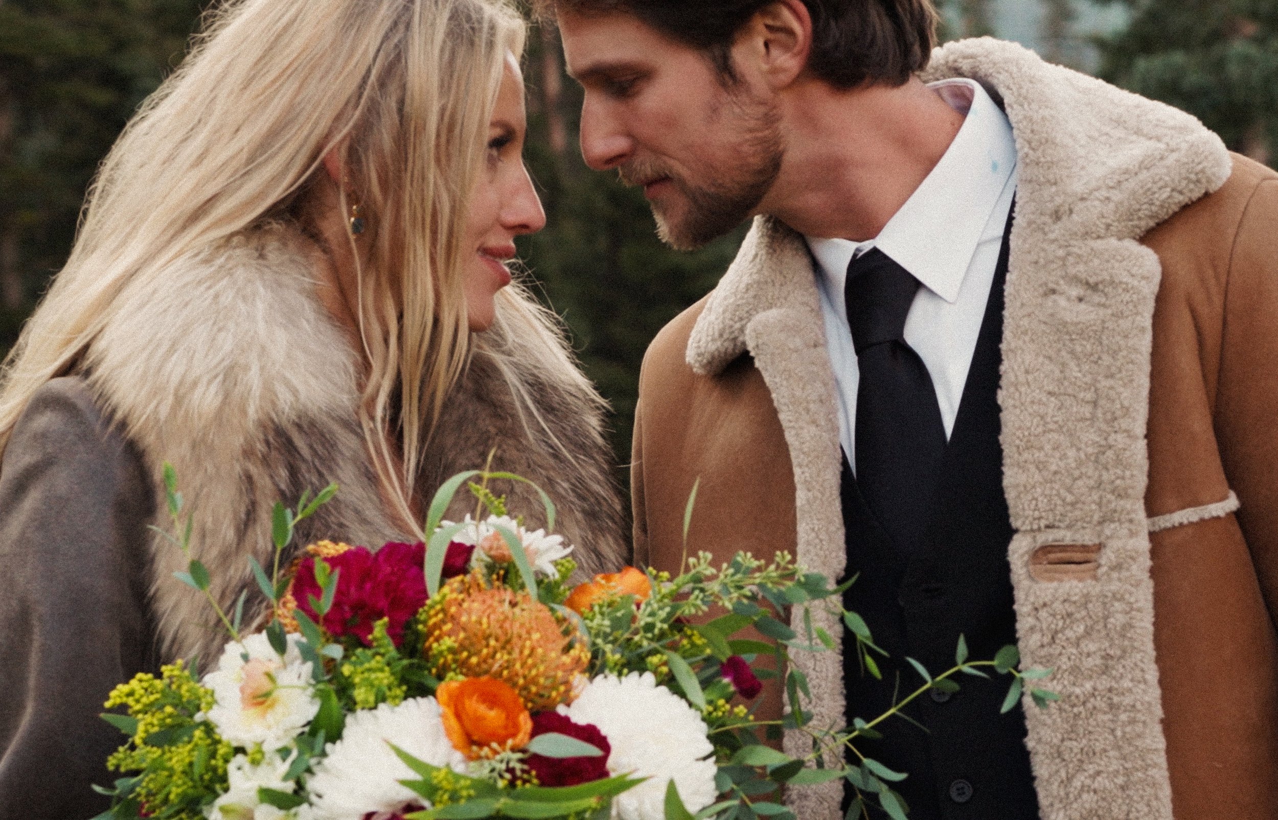 A man and woman face each other close up, outdoors with greenery in the background. The woman holds a colorful bouquet of flowers, and they appear to be sharing an intimate moment.