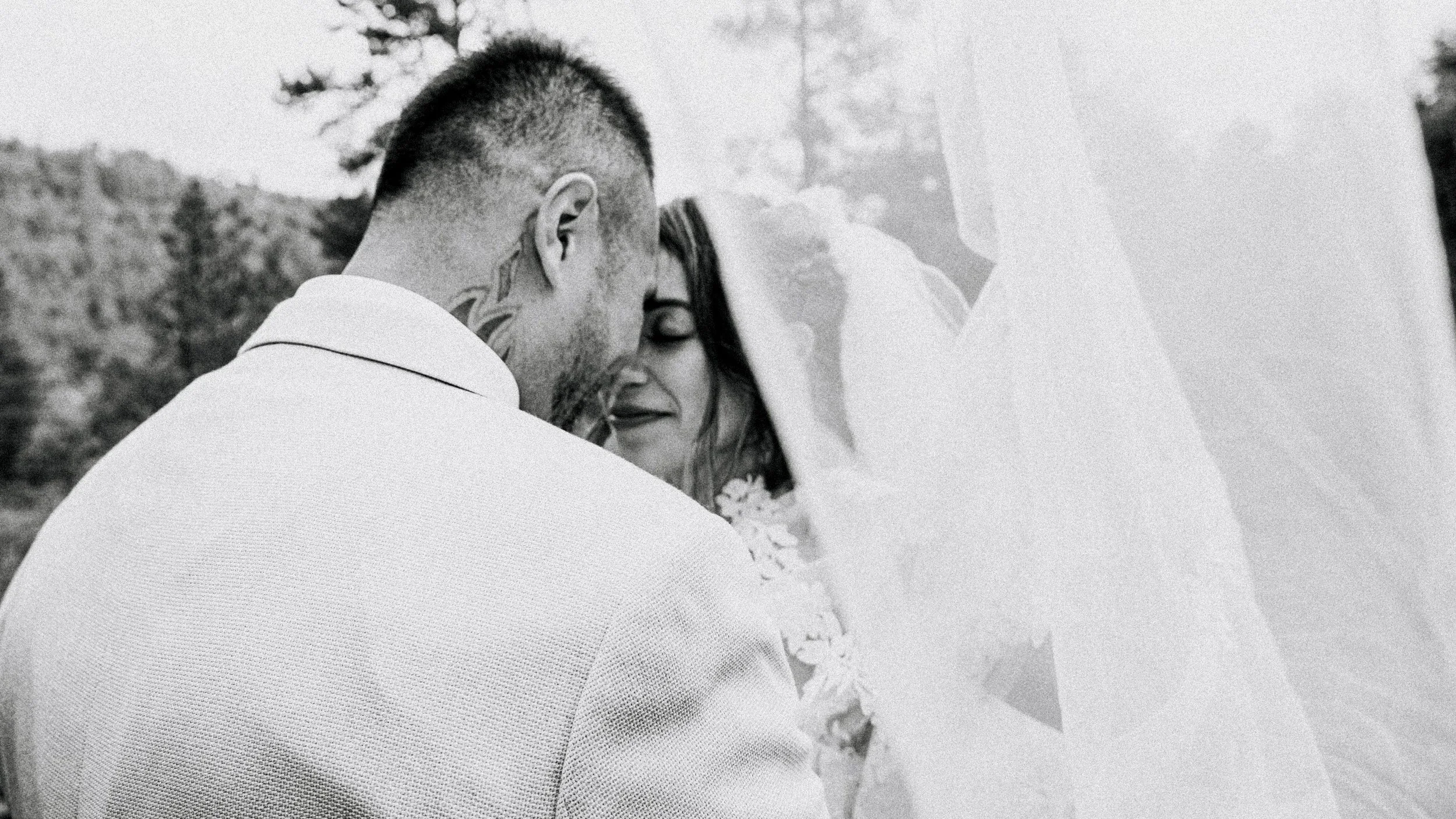 A black and white photo of a couple on their wedding day, with their foreheads touching, eyes closed, sharing an intimate moment outdoors. The bride is wearing a veil and a floral accessory, and the groom has tattoos on his neck and is wearing a white blazer.