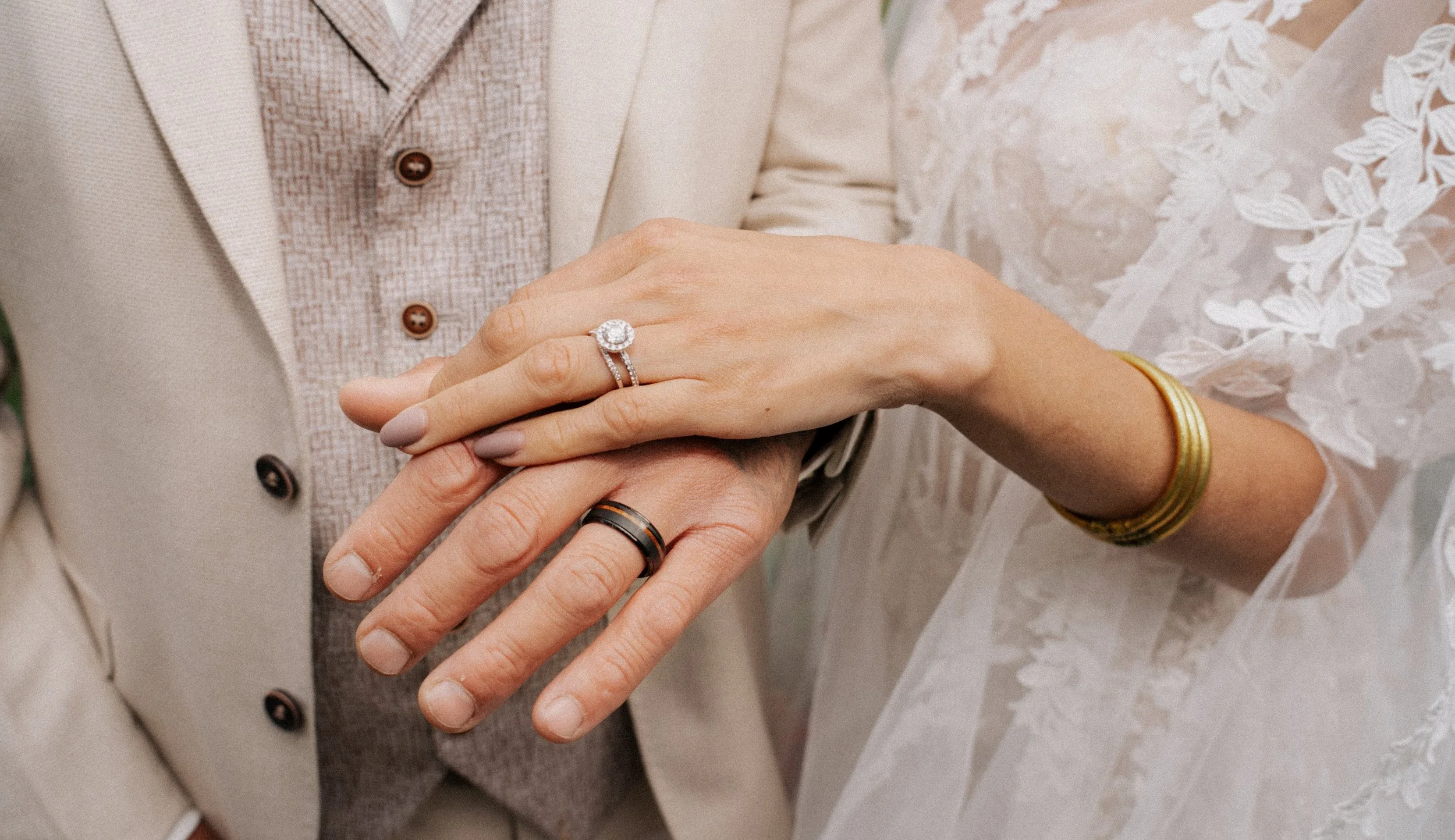 Close-up of a couple's hands showing wedding rings, with the bride wearing gold bangles and the groom wearing a black band, on their wedding day.