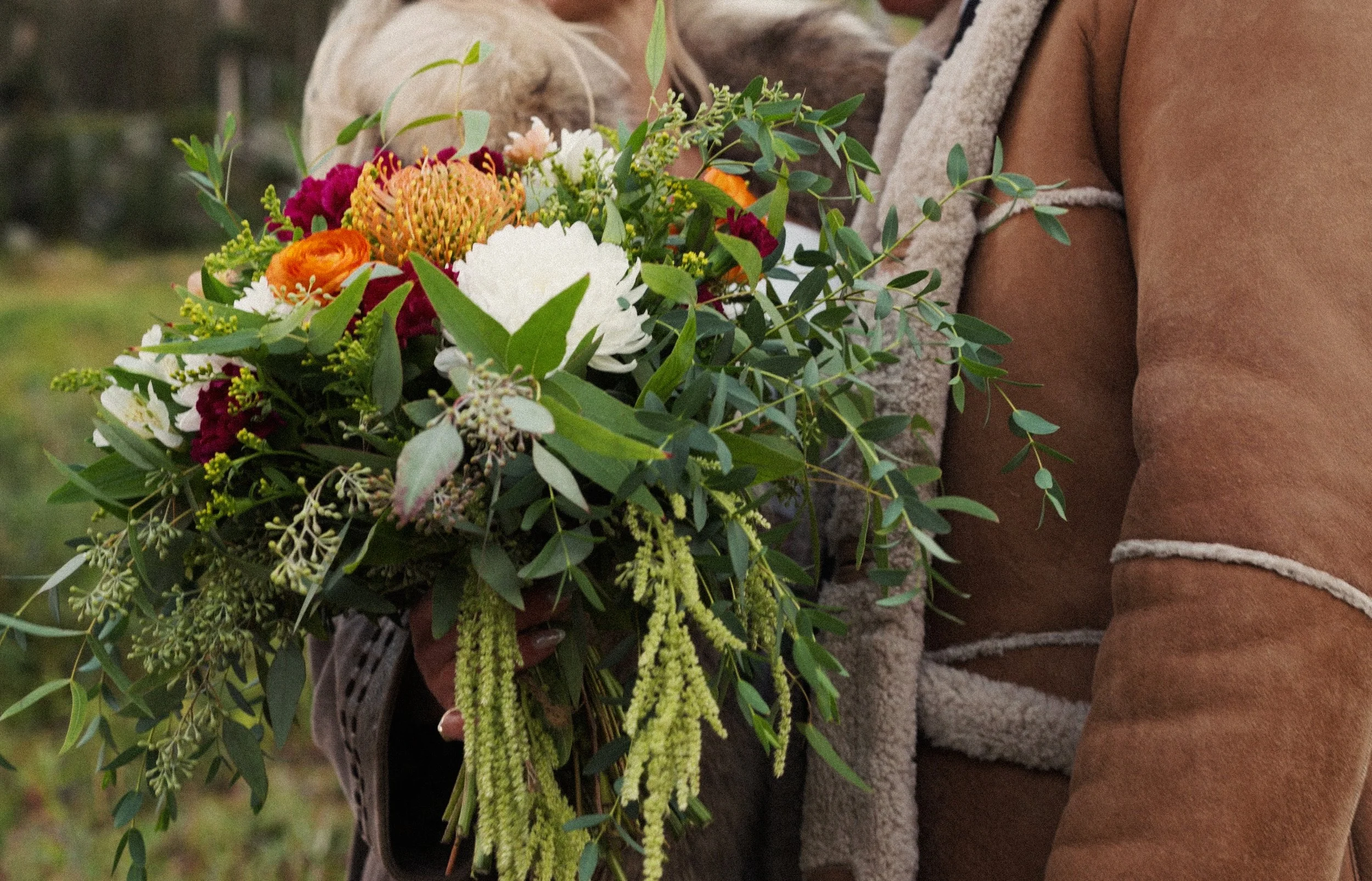 Person holding a bouquet of colorful flowers outdoors, including white, orange, and burgundy blooms with greenery, in a natural setting.