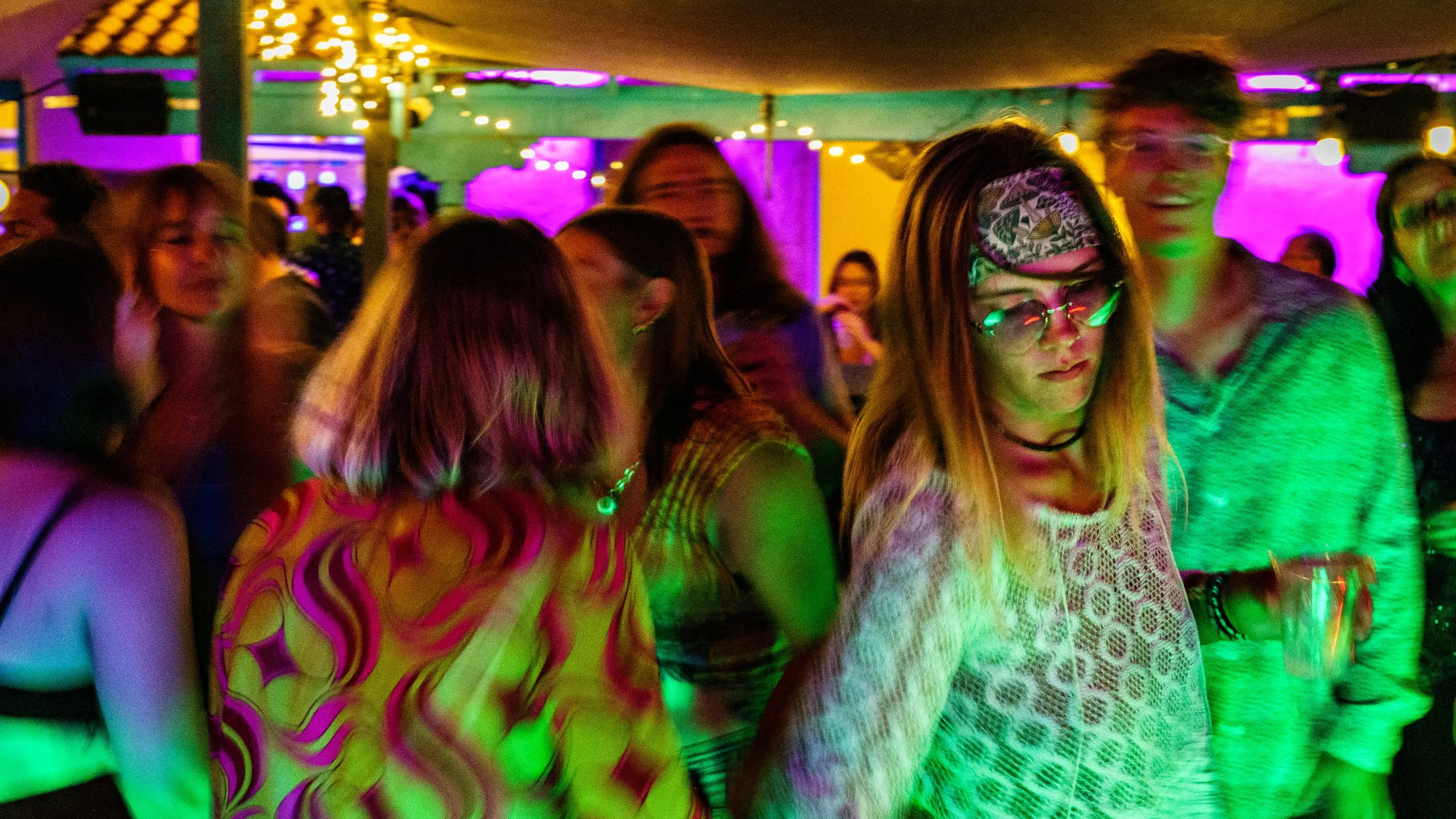 Group of people dancing and socializing at a party or nightclub with colorful lighting and a lively atmosphere.