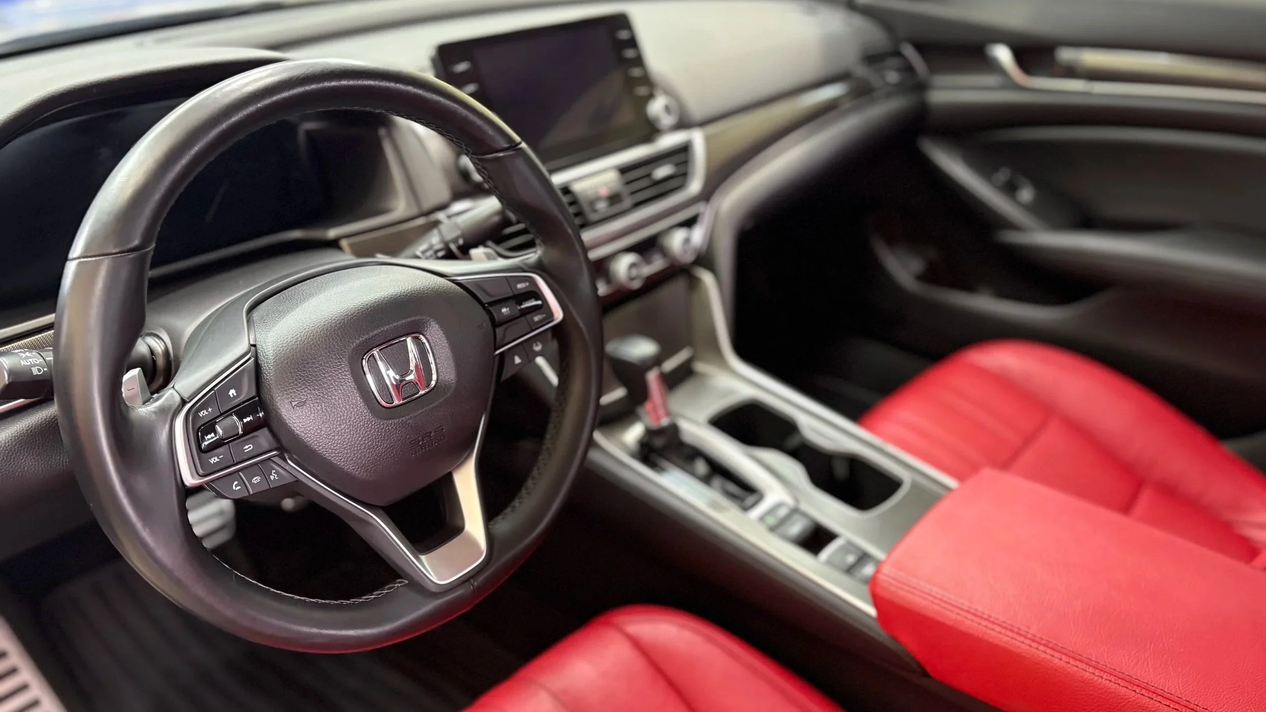 Close-up of a Honda car interior showing a leather-wrapped steering wheel with Honda logo, red leather seats, and the center console with gear shifter.