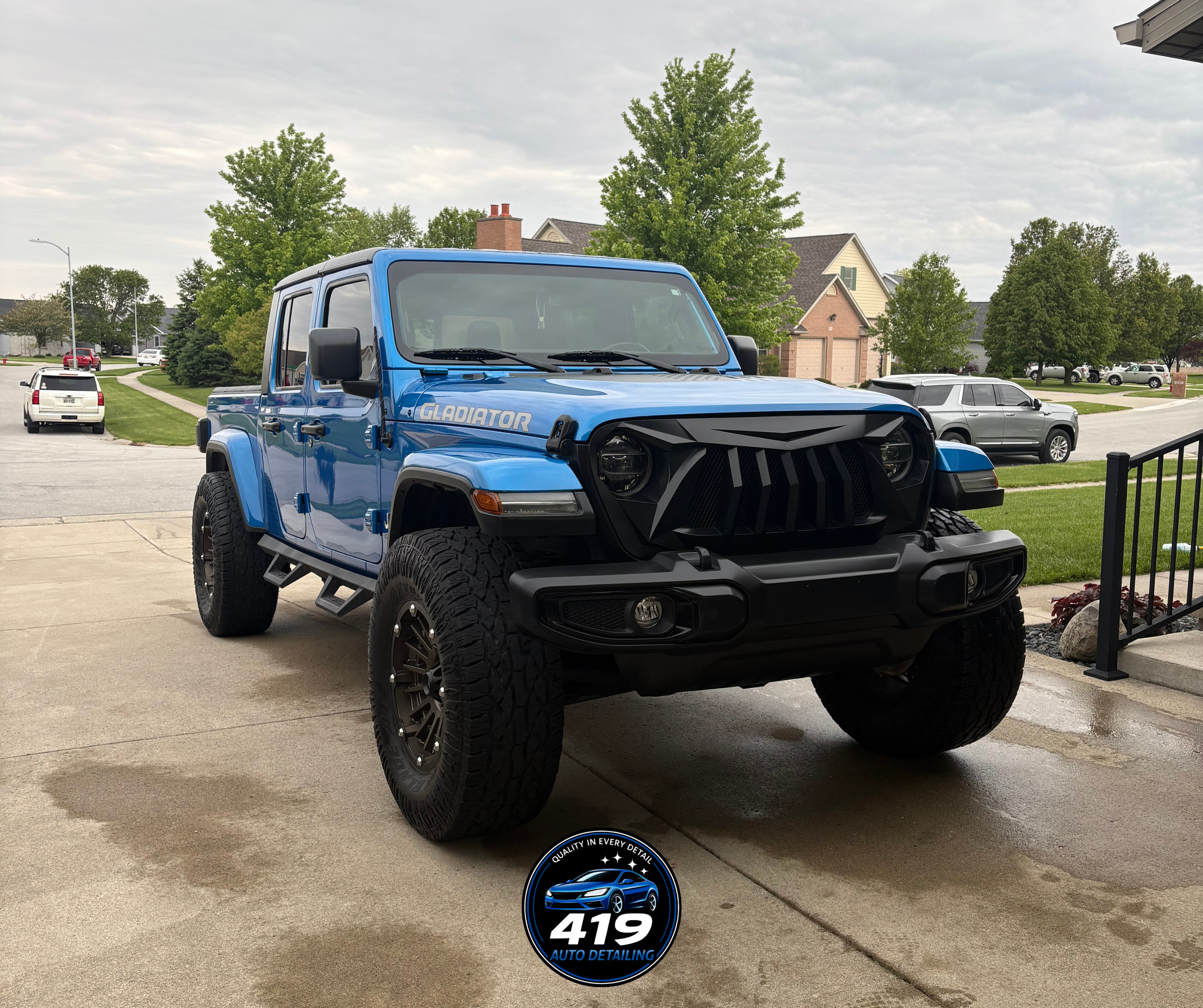 Blue Jeep Gladiator parked in a driveway with a residential neighborhood in the background, including houses, trees, and other parked cars.