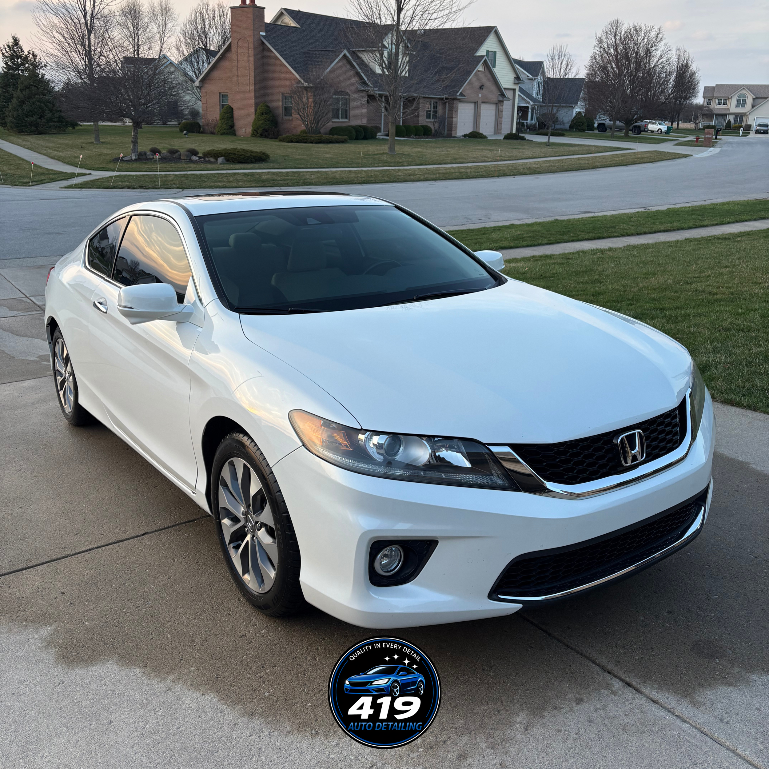 A white Honda sedan parked on a driveway with a suburban neighborhood in the background, including houses, trees, and a street.