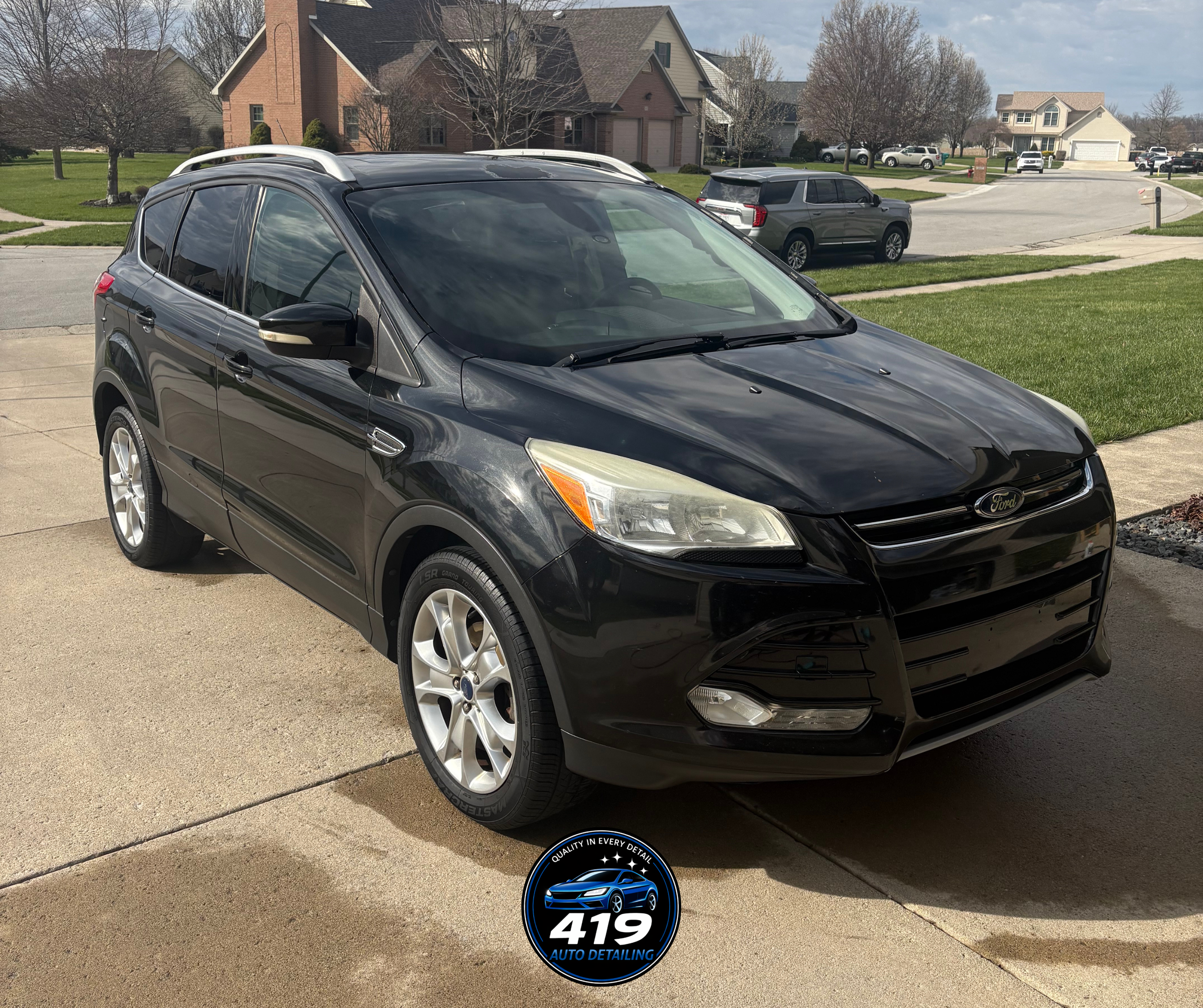 A black Ford SUV parked on a driveway in a suburban neighborhood with houses, green lawns, trees, and a clear sky in the background.