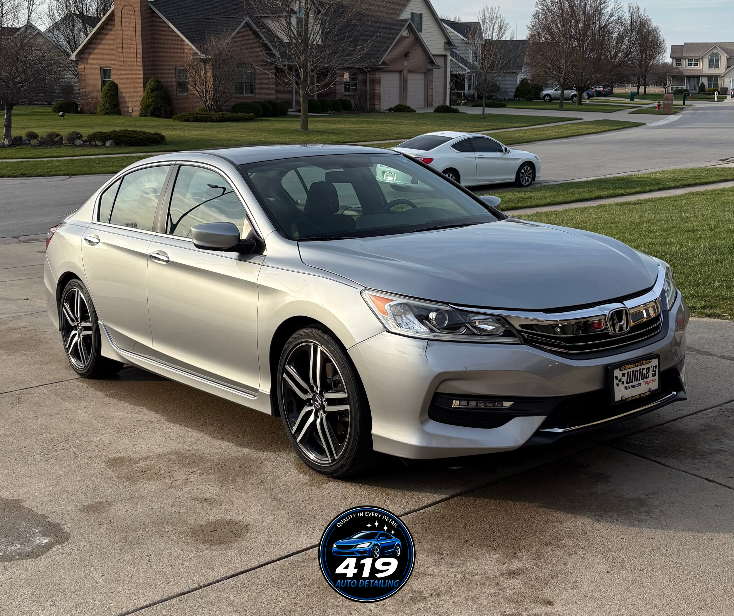 A silver Honda sedan parked on a driveway with a house, trees, and a second white car in the background on a suburban street.