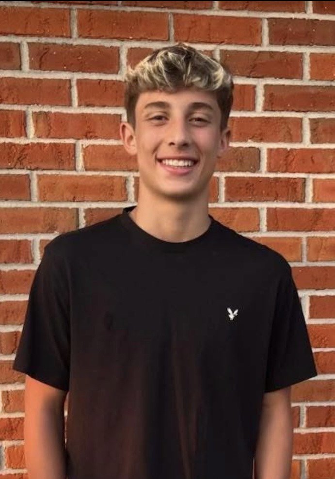 A young man with light brown hair and a blonde highlight, smiling, standing in front of a brick wall. He is wearing a black t-shirt with a small white eagle logo on the chest.