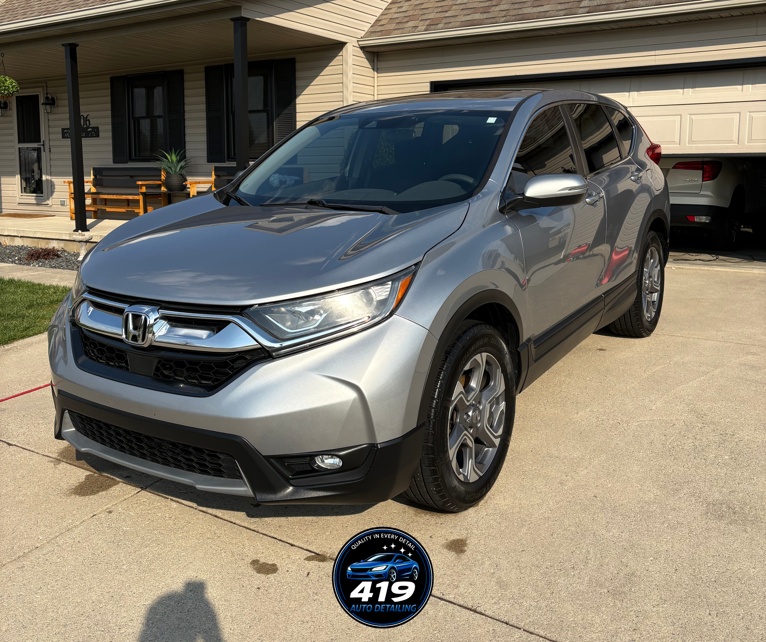Silver Honda SUV parked in driveway in front of a house with beige siding, black shutters, and a garage door. A black and blue auto detailing sticker is on the driveway.