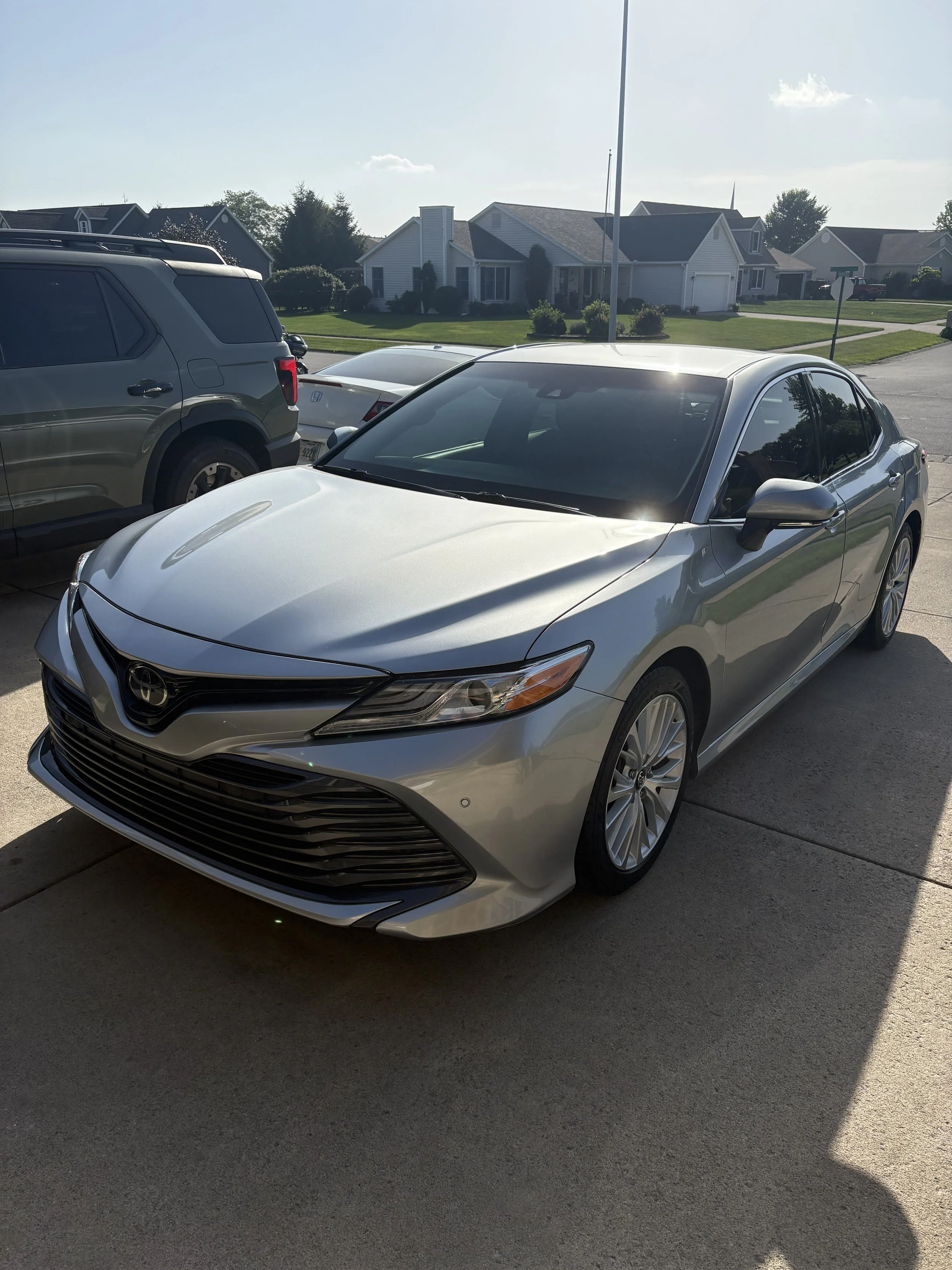 A silver Toyota Camry parked on a driveway in front of a suburban house with green lawns and neighboring houses in the background, during daytime with clear skies.