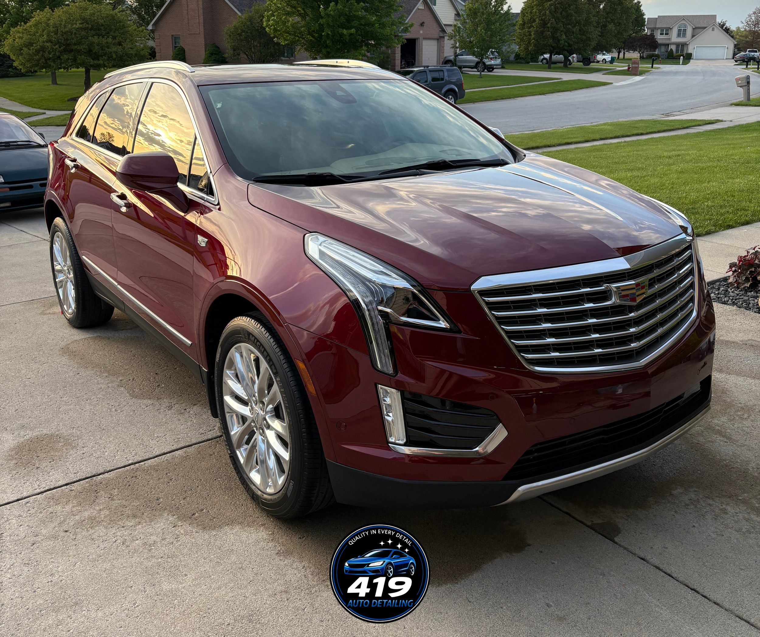 Red Cadillac SUV parked on driveway in front of suburban residential neighborhood with trees, houses, and street visible in background at sunset.