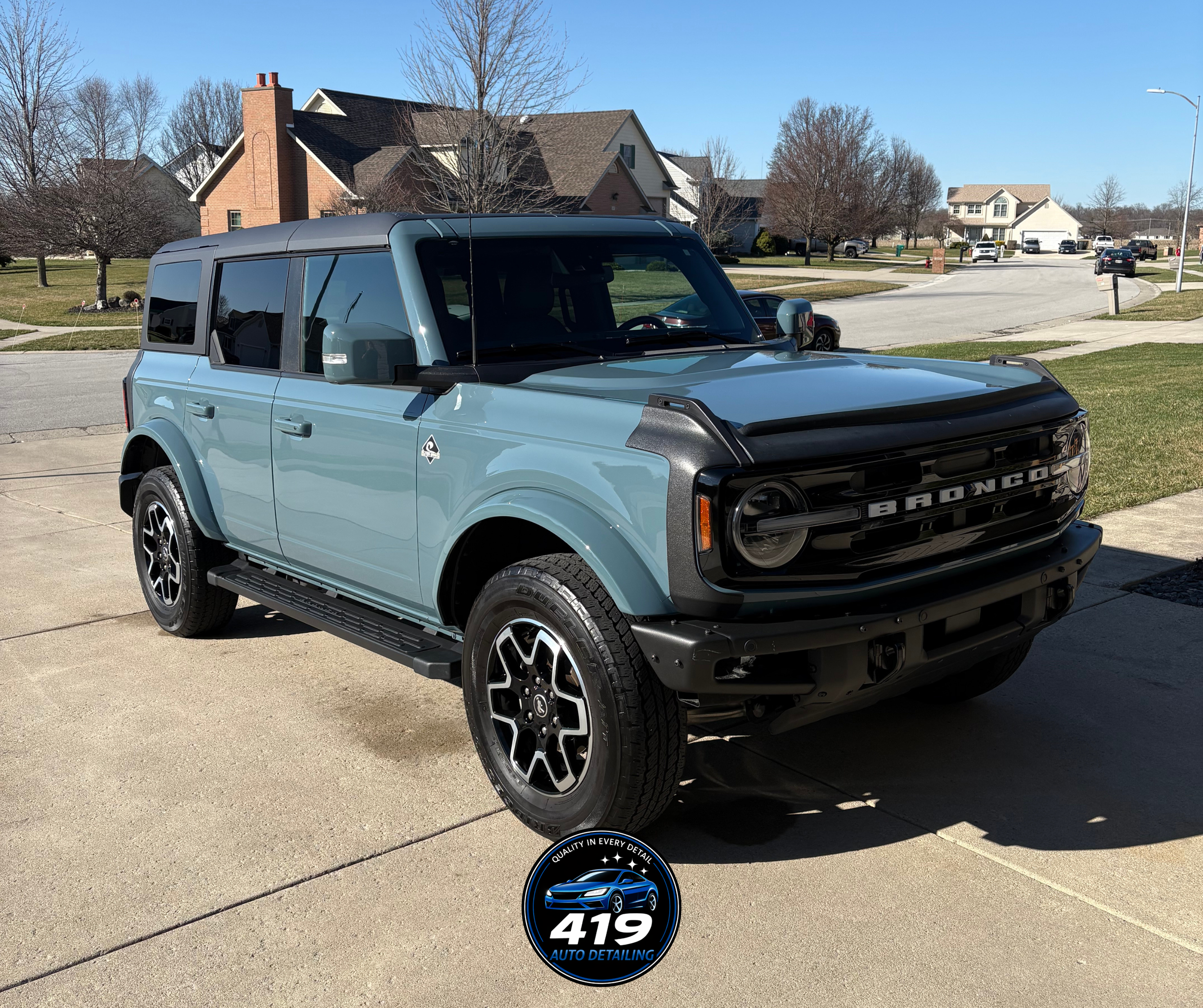 Light blue Ford Bronco parked in a driveway with a black grille and trim, and black and silver wheels, with houses and trees in the background.