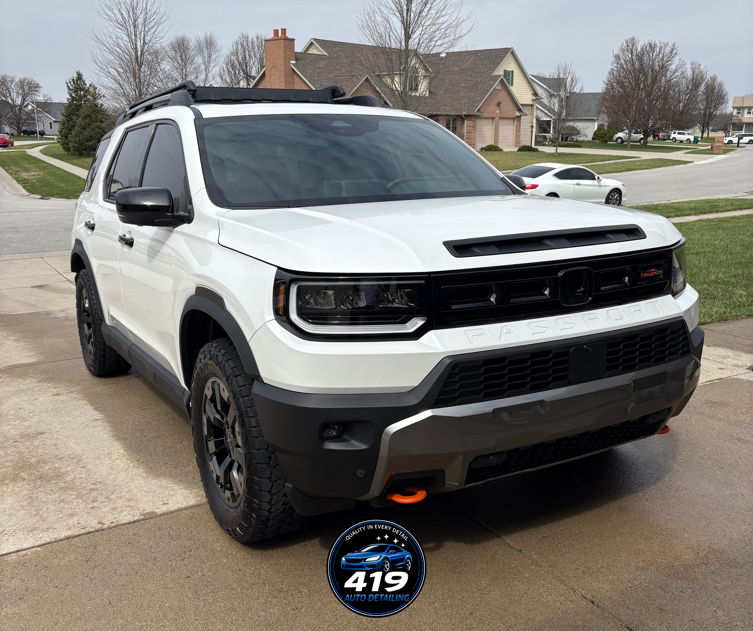 White Honda Passport SUV parked on driveway in a neighborhood with houses and trees in the background, featuring black off-road tires, black side mirrors, a black grille, and a small orange winch hook at the front. Logo of 419 Auto Detailing is at th