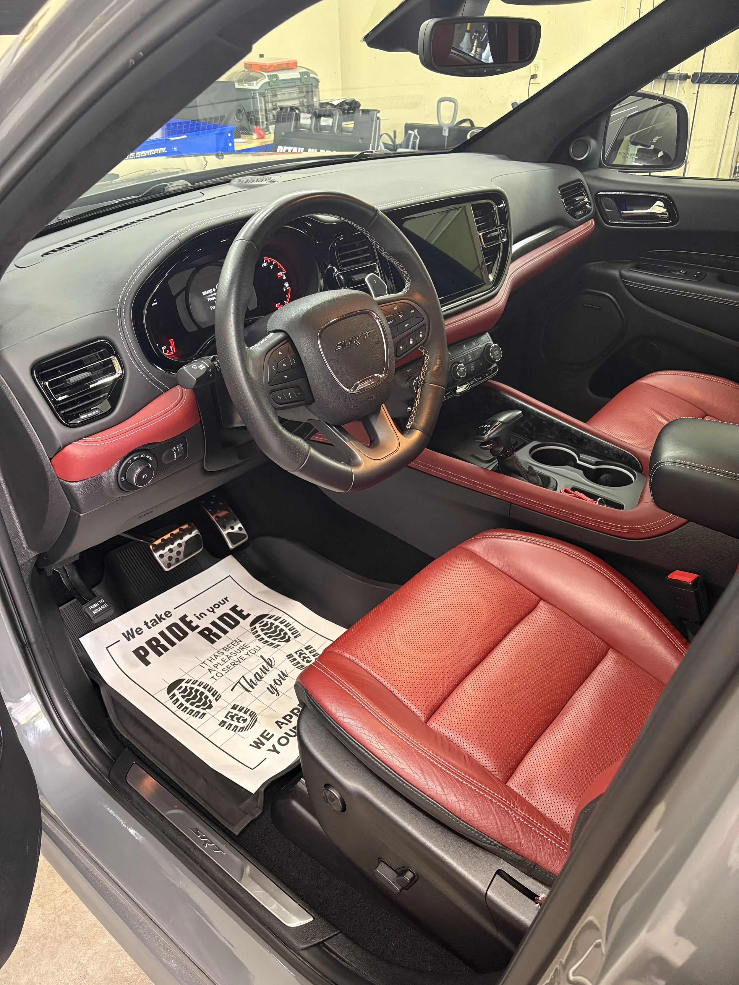 The interior of a sports car with red leather seats, a black dashboard, and a steering wheel with the SRT logo, showing pedals and a floor mat with promotional text.
