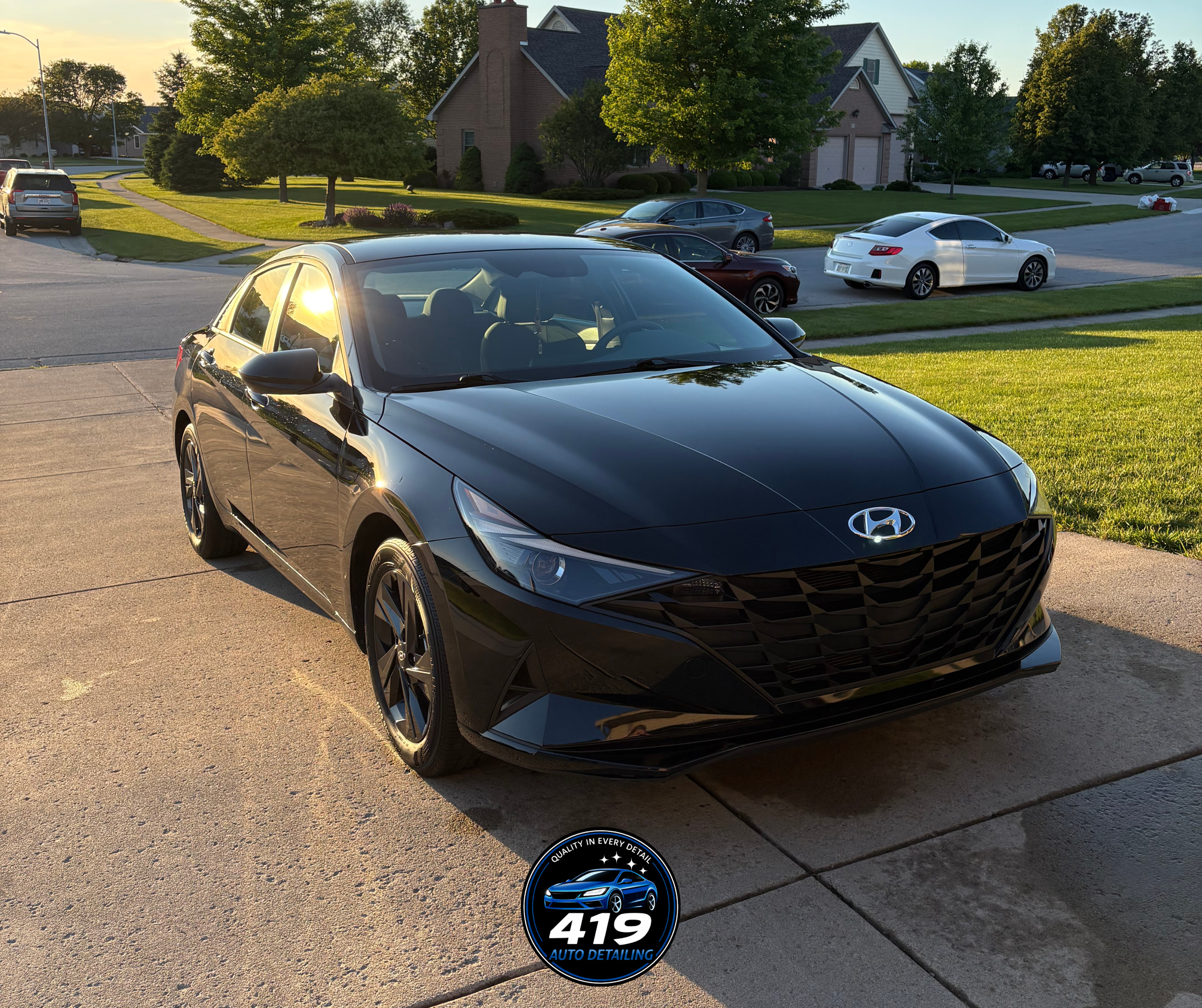 A black Hyundai sedan parked on a driveway in a suburban neighborhood during sunset, with other cars, houses, trees, and a well-manicured lawn visible in the background.