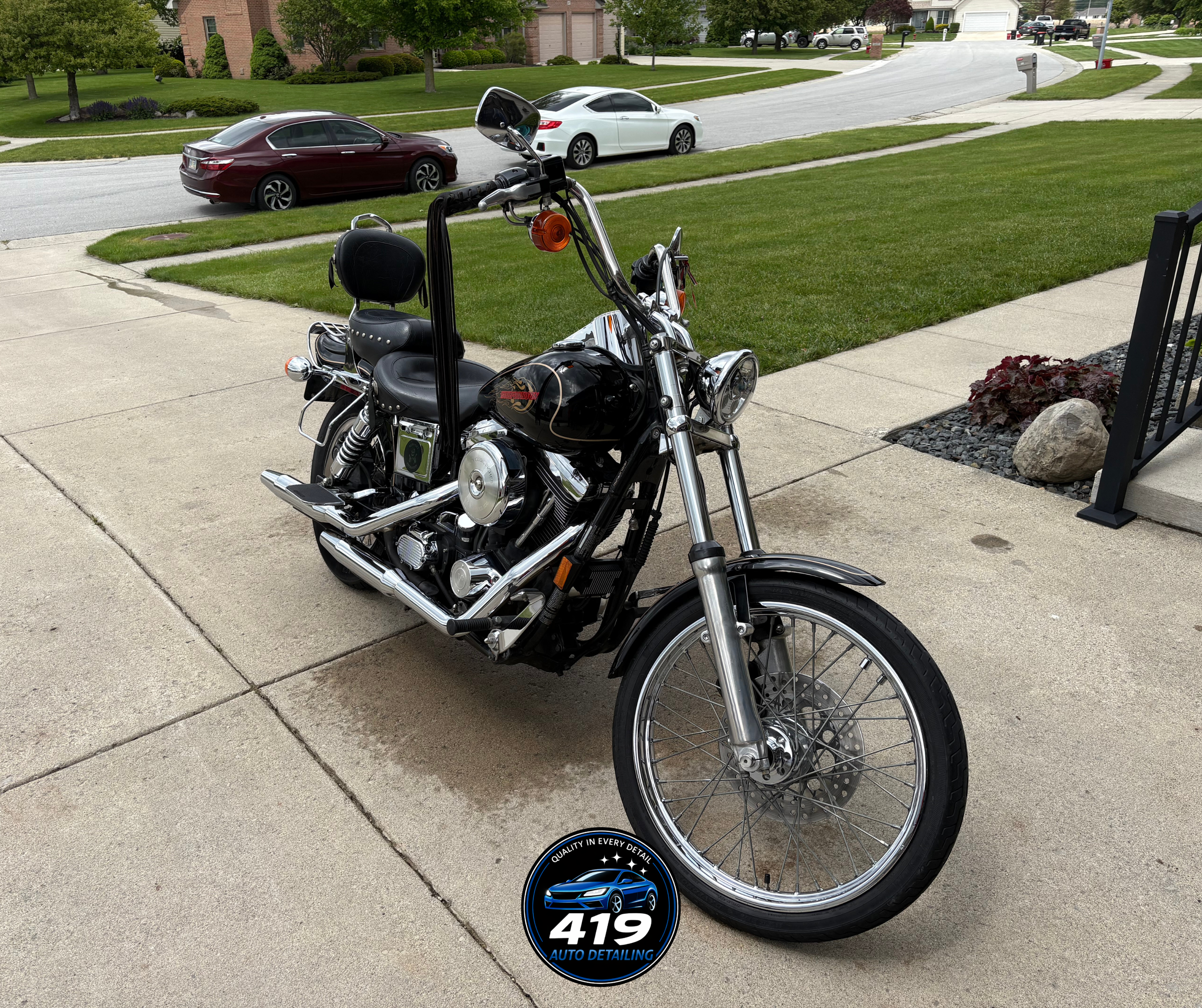 A black motorcycle parked on a sidewalk in front of a green lawn. In the background, there are three parked cars on a street, trees, houses, and a mailbox. The motorcycle has a Harley Davidson logo on the fuel tank and a round headlight. There is a c