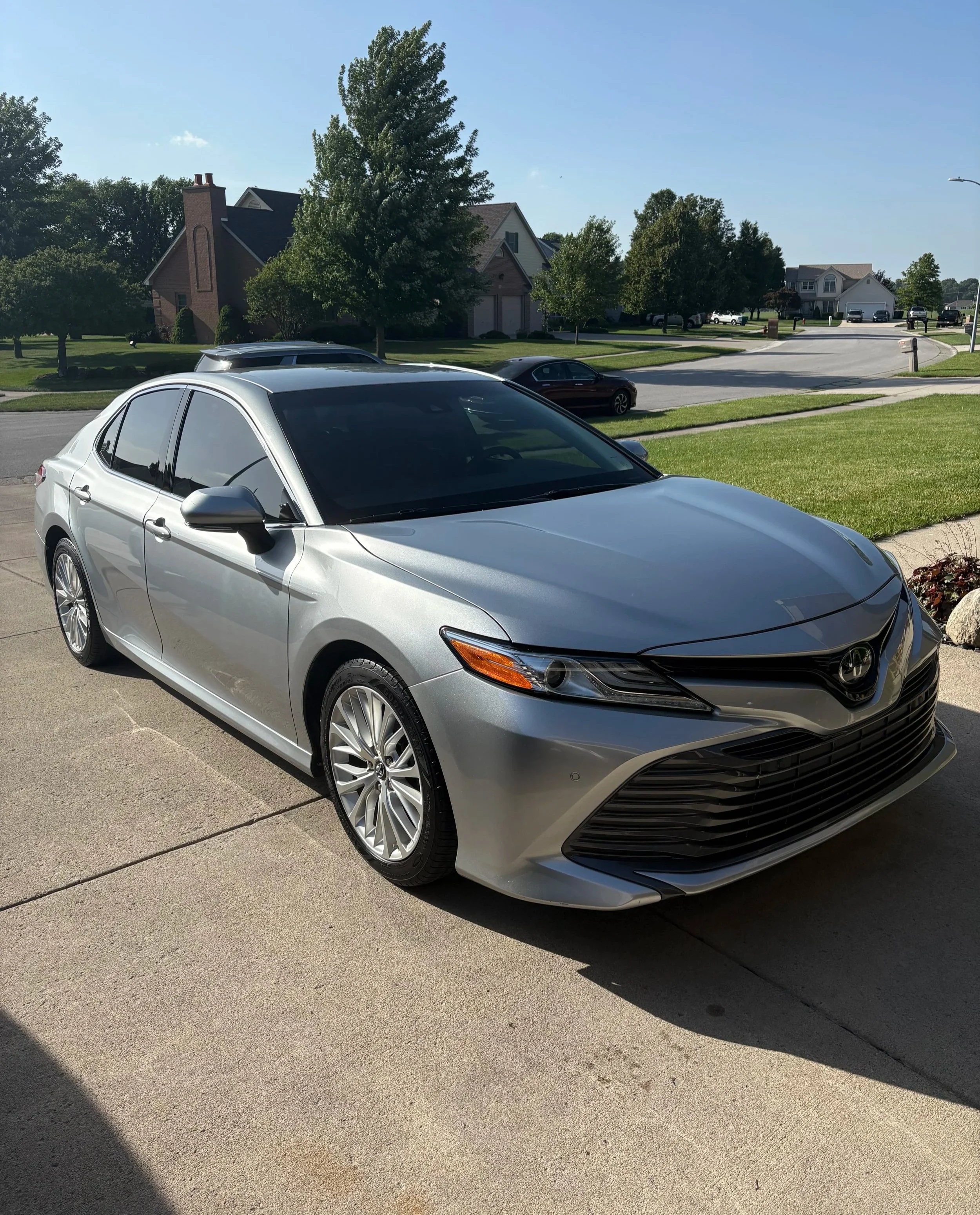 A silver sedan parked on a driveway in a suburban neighborhood with green lawns and houses in the background under a clear blue sky.