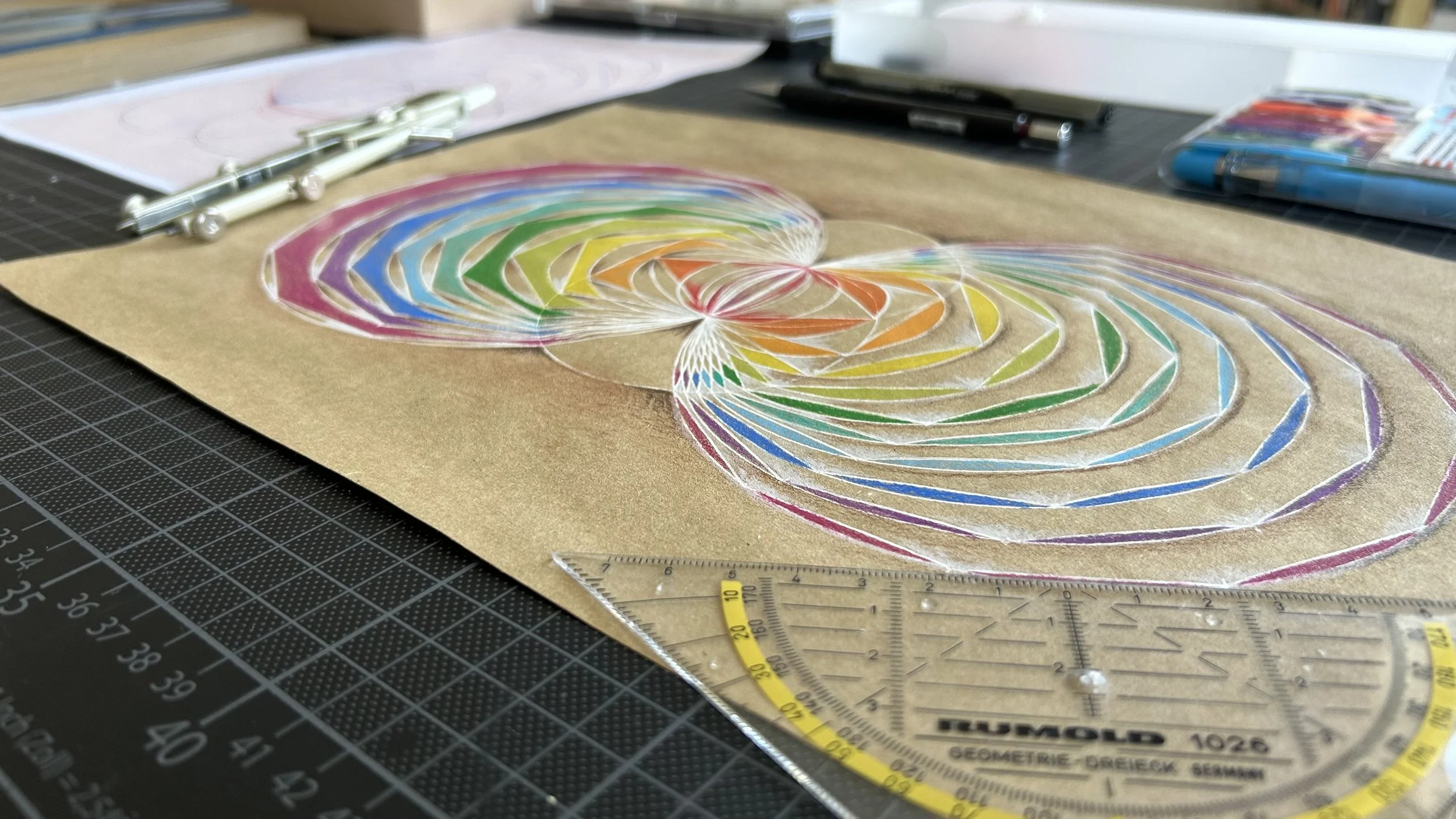 Kunstprojekt mit bunten, spiralförmigen Mustern aus Krepppapier auf einem braunen Untergrund. Es liegen Werkzeuge wie ein Geodreieck, Marker und Markerboxen im Hintergrund.