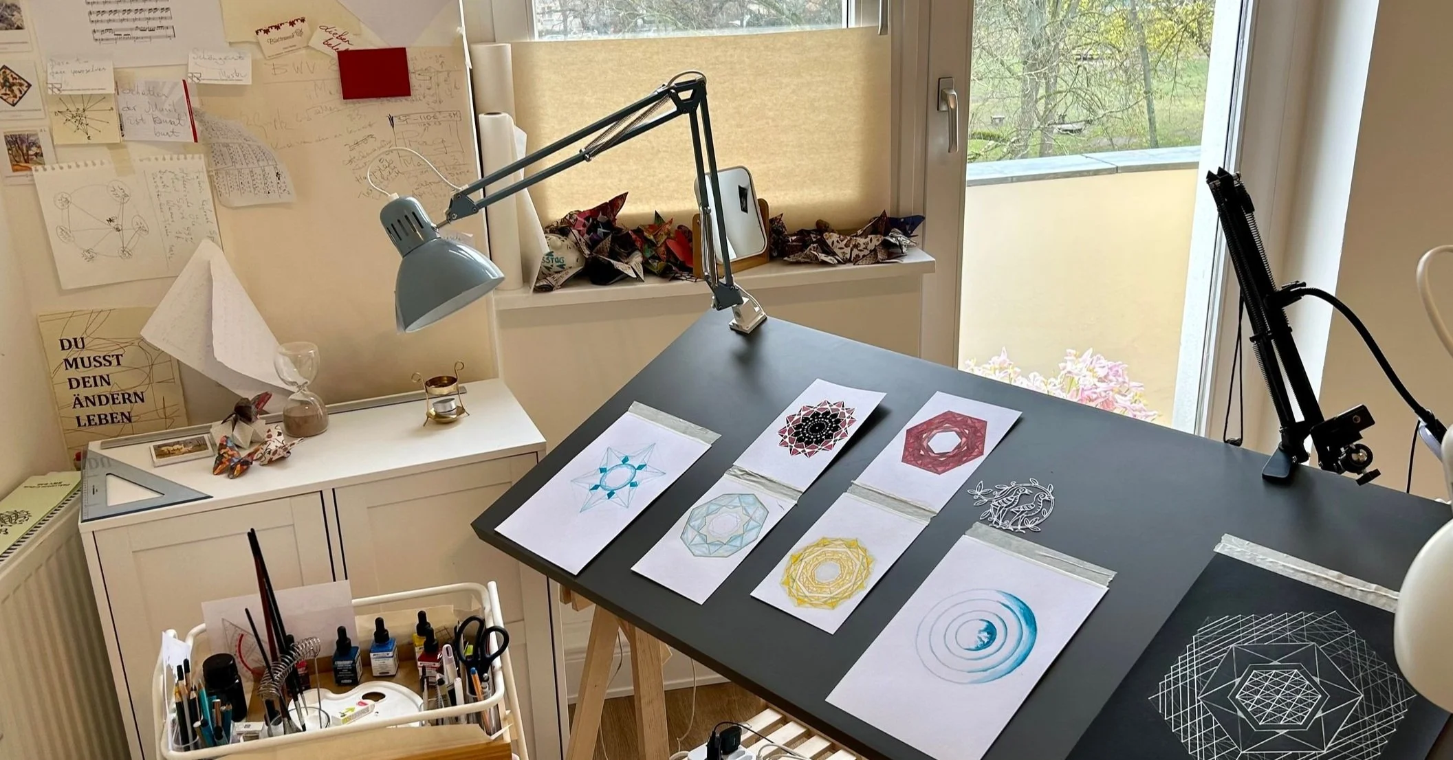 Ein Arbeitsplatz mit Zeichen- und Designmaterialien, darunter geometrische Muster auf Papier, Zeichenutensilien in einer Schale, eine Tischlampe und ein schwarz gedeckter Tisch mit zusammengelegten Entwürfen, an einer Fensterseite mit Blick nach draußen.