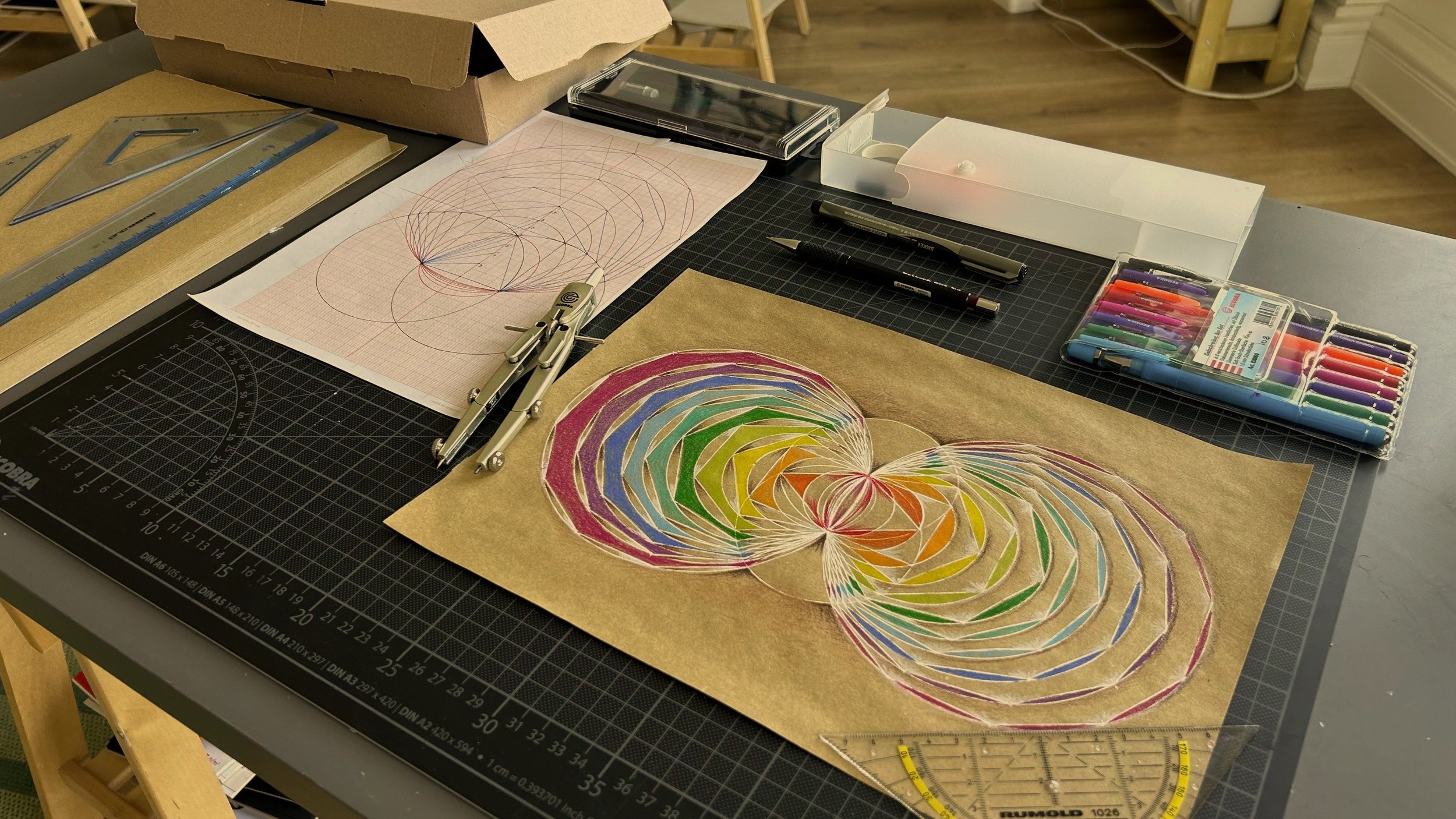 Arbeitsplatz mit Zeichenmaterialien, einer colorierten Zeichnung, einem Linien- und Kreisdiagramm, technischen Werkzeugen, einem Lineal, einem Stift und bunten Filzstiften auf einer schwarzen Schneidematte.