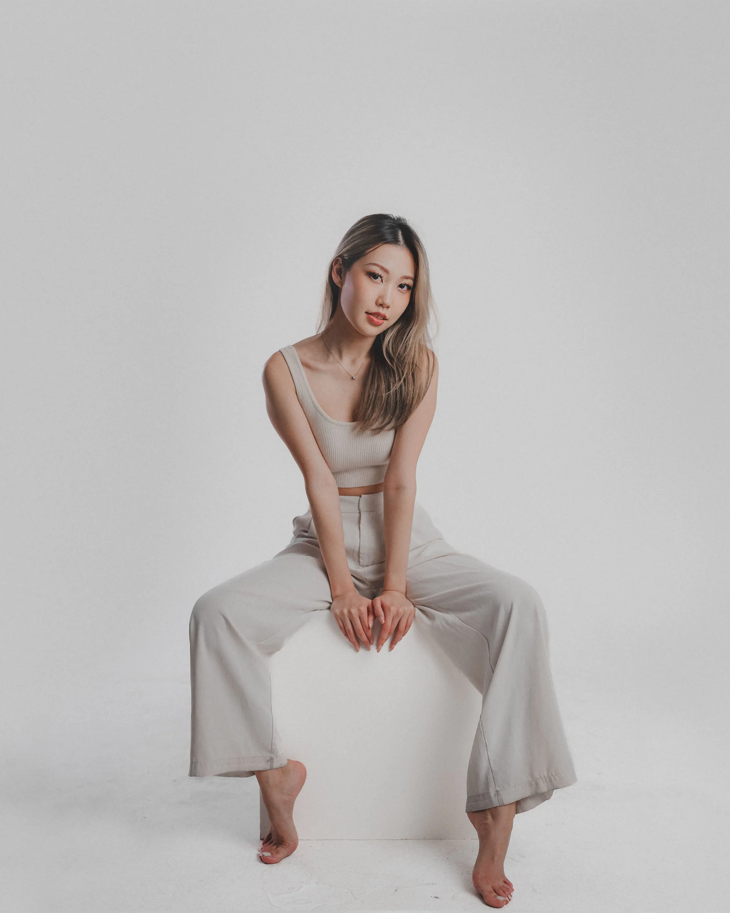A woman sitting on a white box, resting her foot on the floor, in front of a plain white background. She has long wavy hair, wears a beige sleeveless top, and loose beige pants.