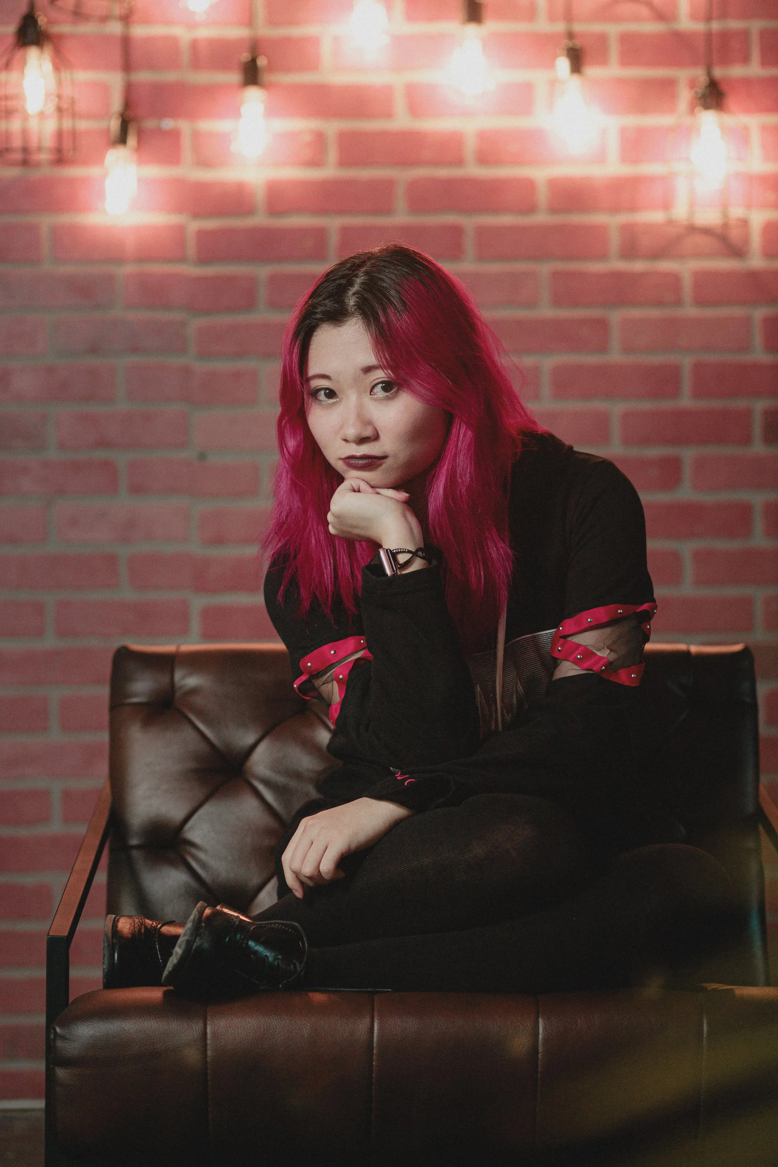 A young woman with pink hair sitting on a dark brown leather couch in front of a red brick wall with hanging Edison bulb lights.