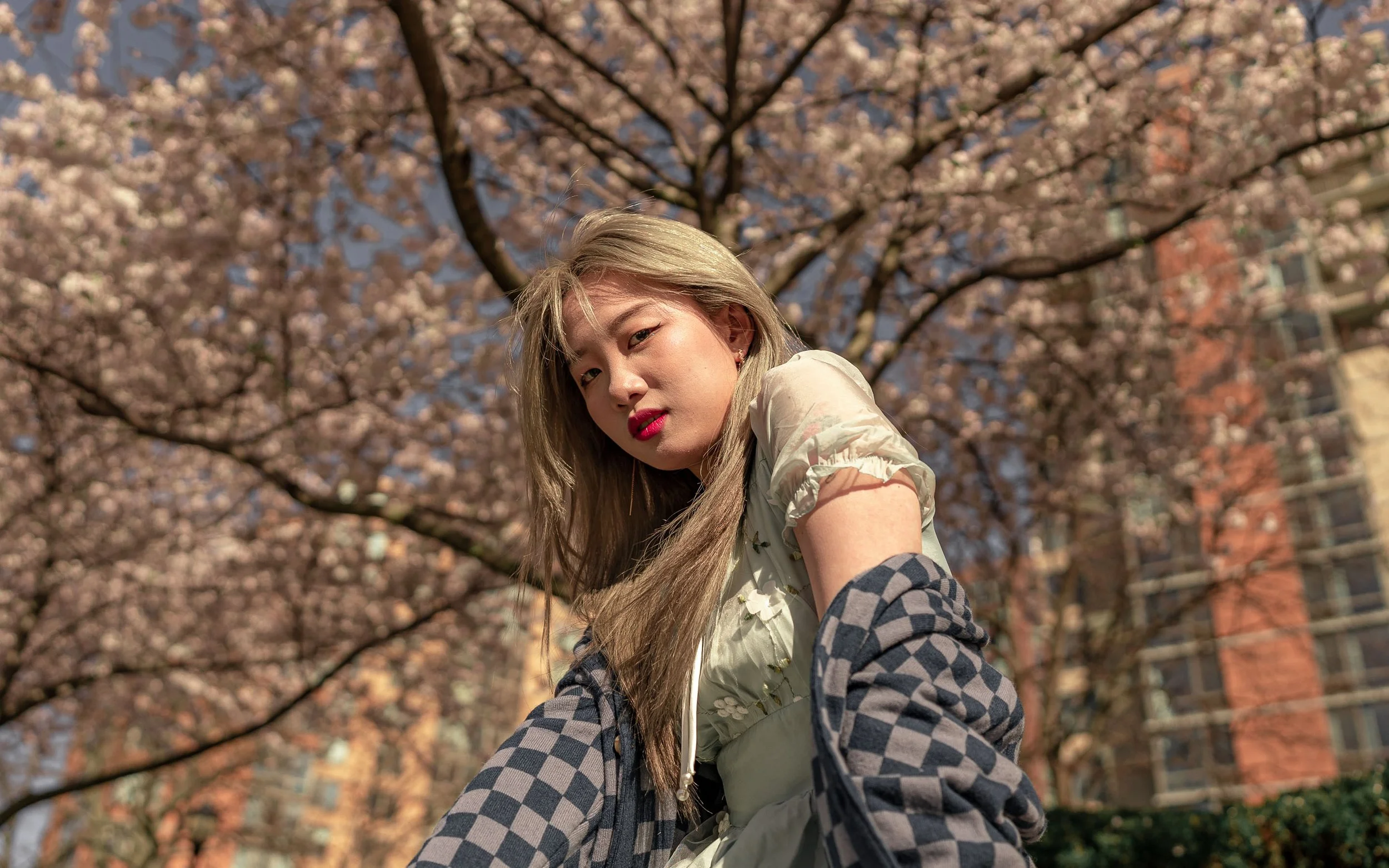 A young woman with long blonde hair and pink lipstick squints and poses outside in front of blooming cherry blossom trees on a sunny day.