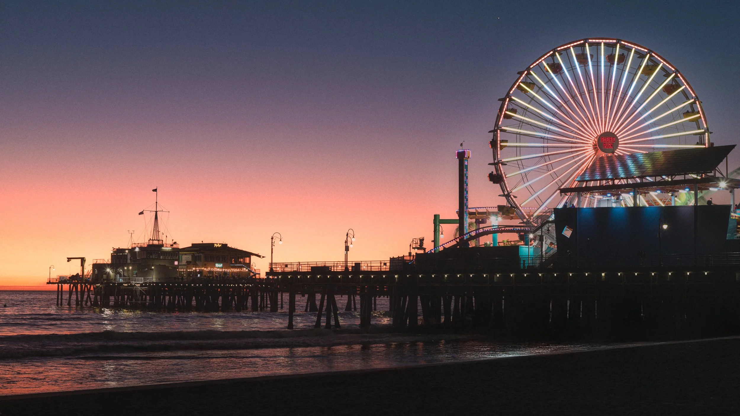 A pier at sunset with a Ferris wheel illuminated with colorful lights in Pacific Park, Santa Monica, California.