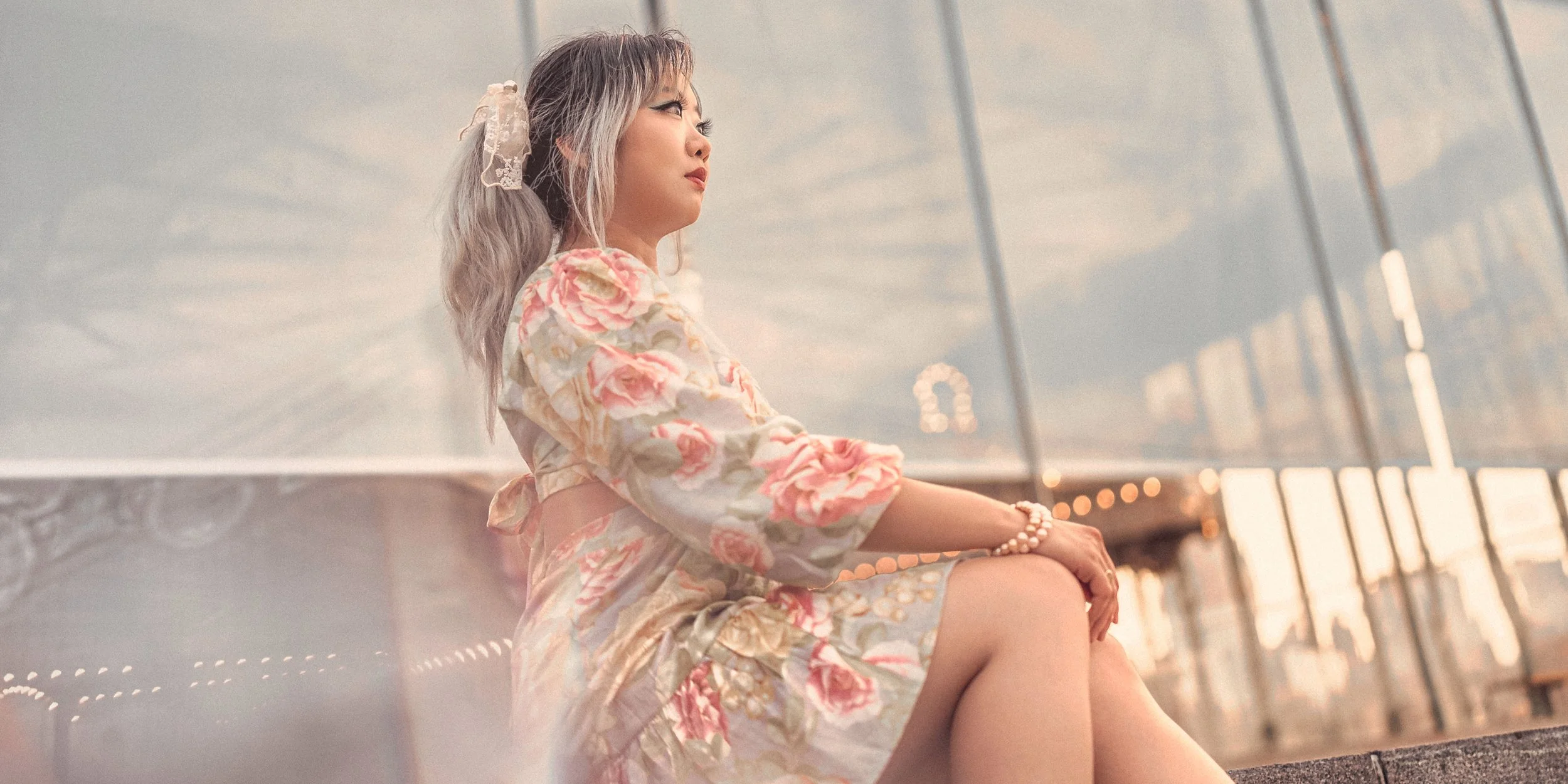 A woman in a floral dress sitting on a bench at an airport terminal with large windows, looking thoughtfully into the distance.