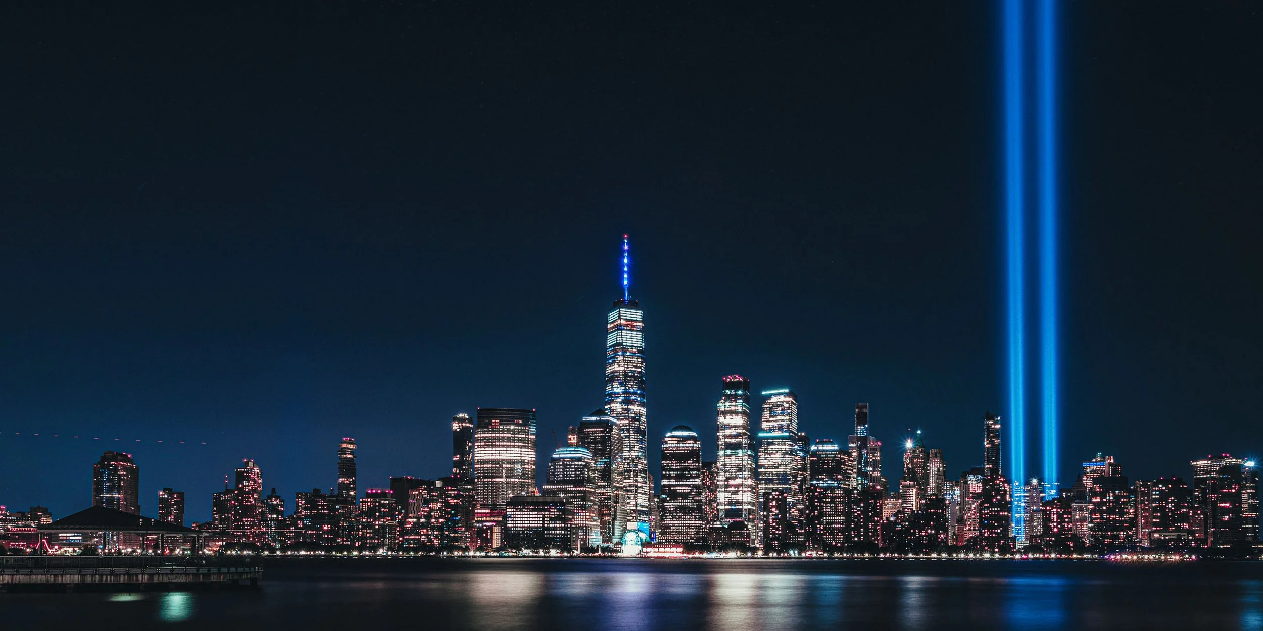 Nighttime city skyline with One World Trade Center, illuminated buildings, and blue light beams rising into the sky reflected in the water.