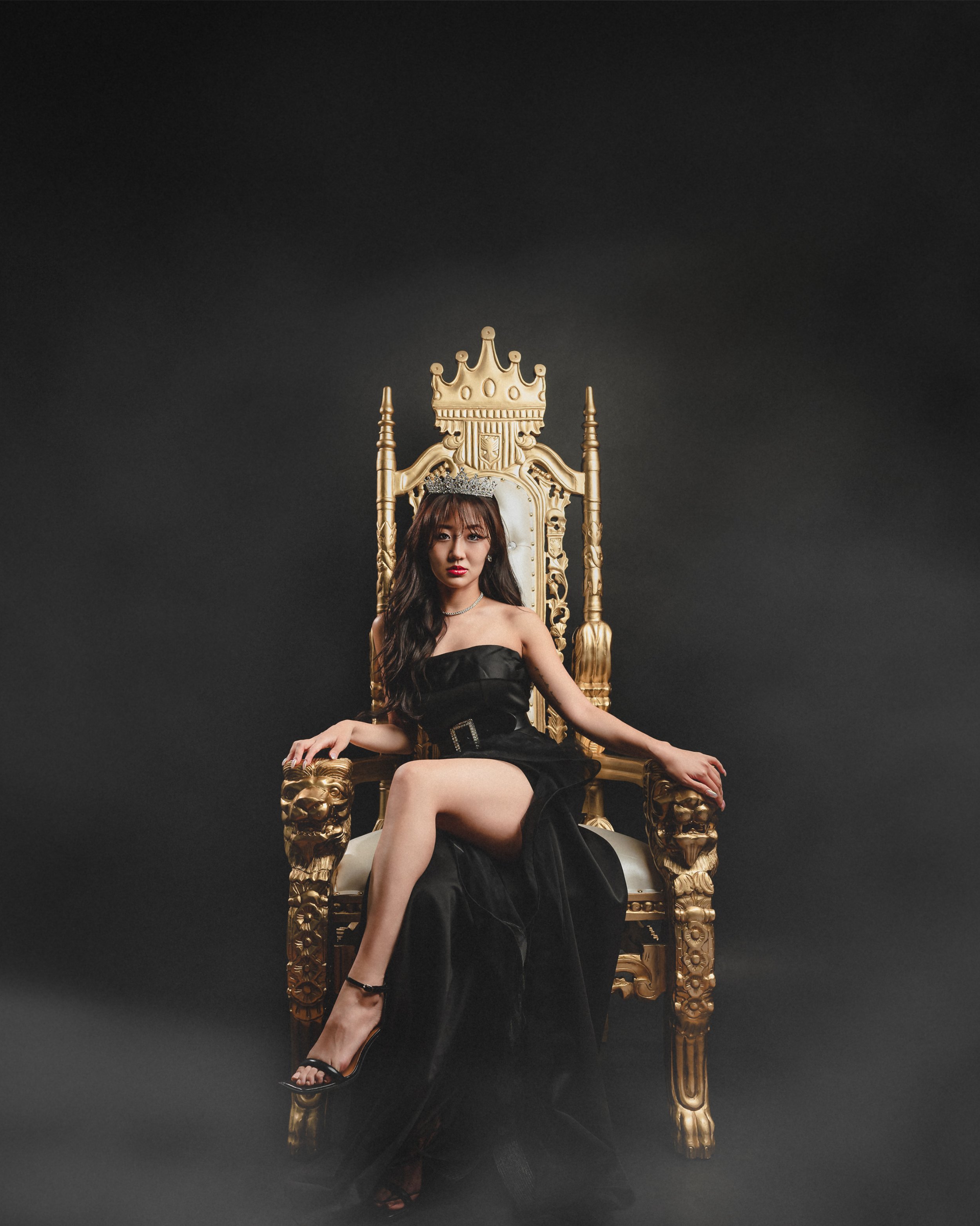 A woman sitting on an ornate gold throne with a crown on her head against a black background, dressed in a black strapless dress with one leg crossed over the other.