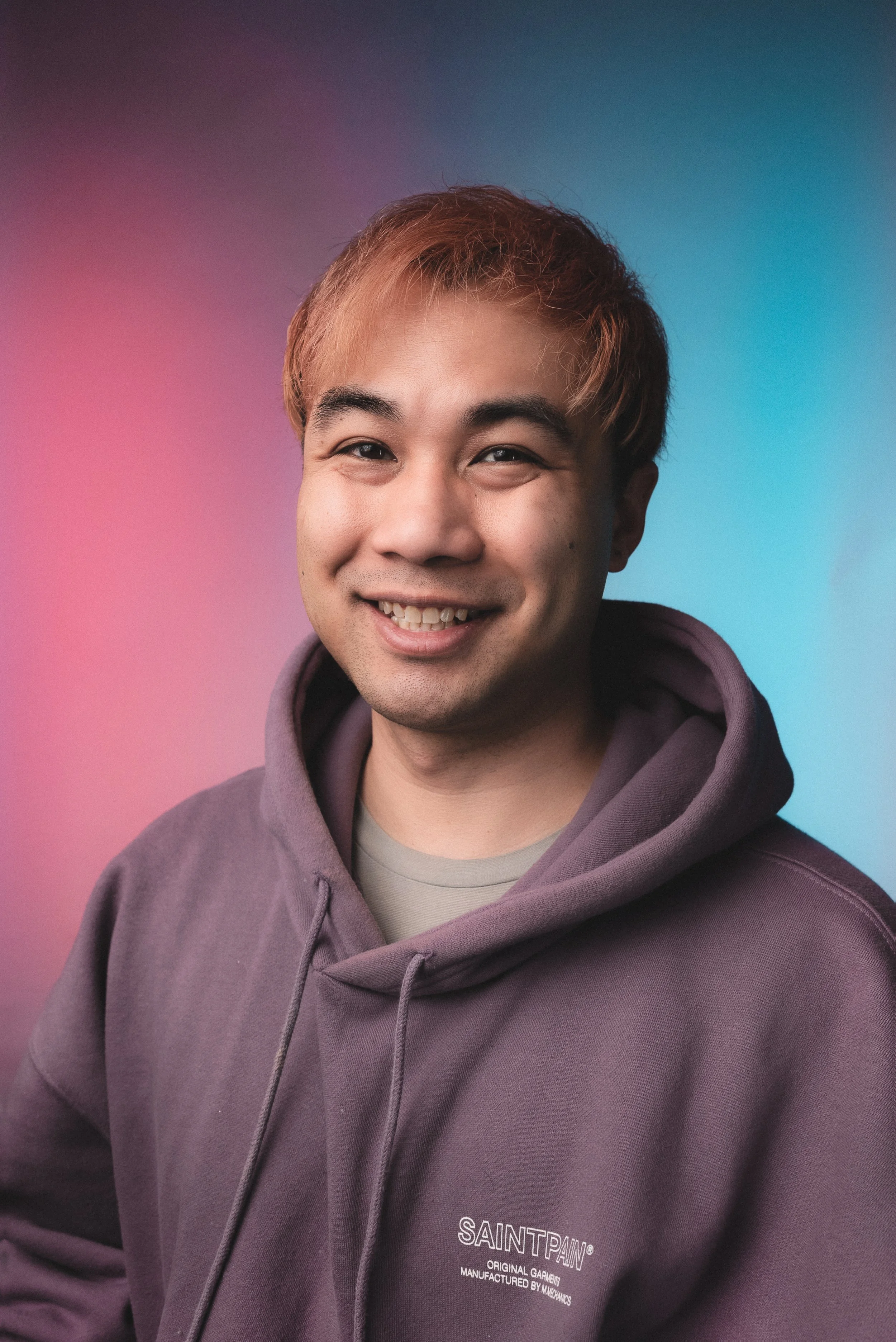 Smiling young man with dark eyebrows, short brown hair, wearing a purple hoodie with white text on the chest, against a colorful gradient background.