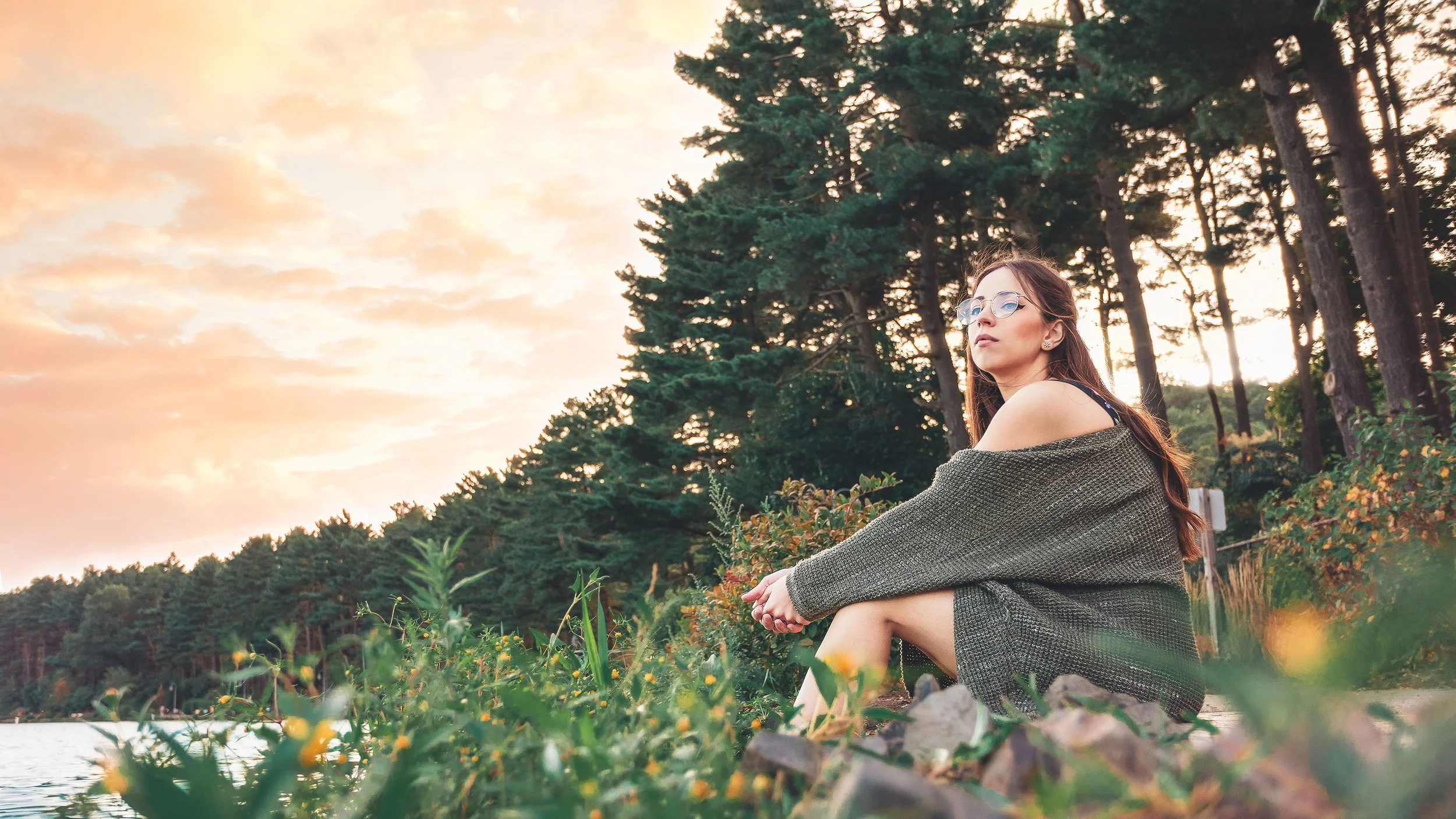 A woman sitting on the rocky shore of a lake during sunset, surrounded by lush green trees, wearing glasses and a green sweater, gazing thoughtfully into the distance.