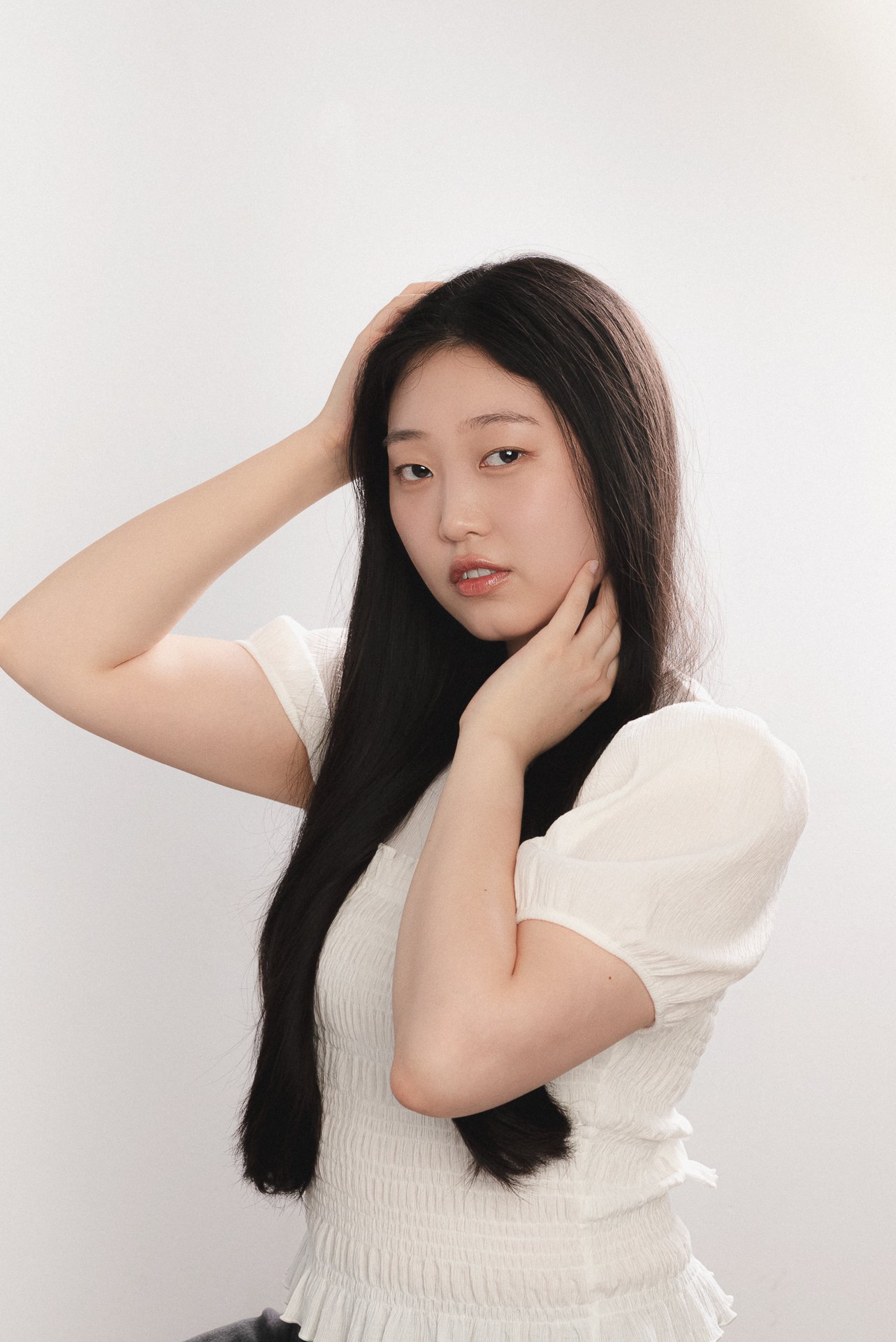 A young woman with long black hair poses against a white background, wearing a white short-sleeved top and a matching skirt, with one hand behind her head and the other lightly touching her neck.