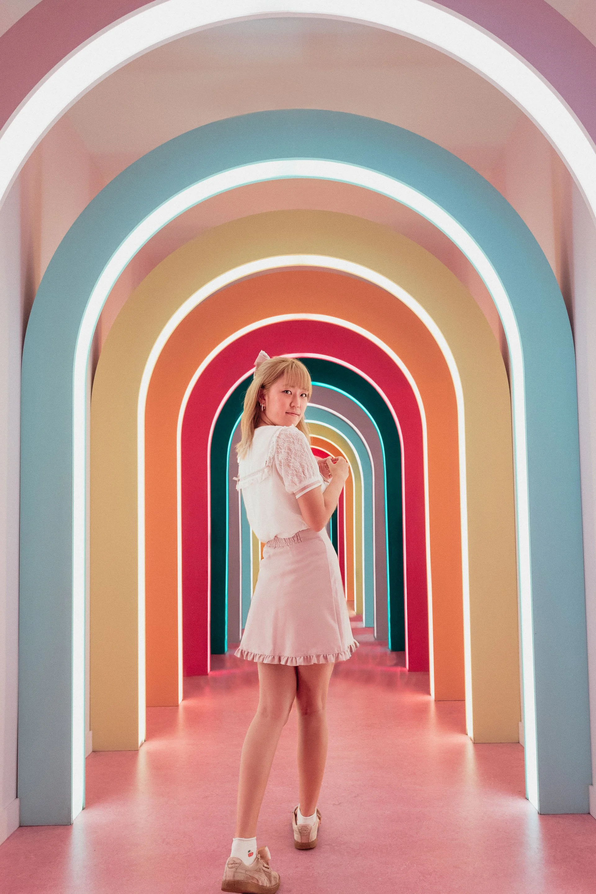 A young woman with blonde hair in a white blouse and pink skirt standing in a colorful, tunnel-like corridor with archways illuminated by multicolored neon lights.