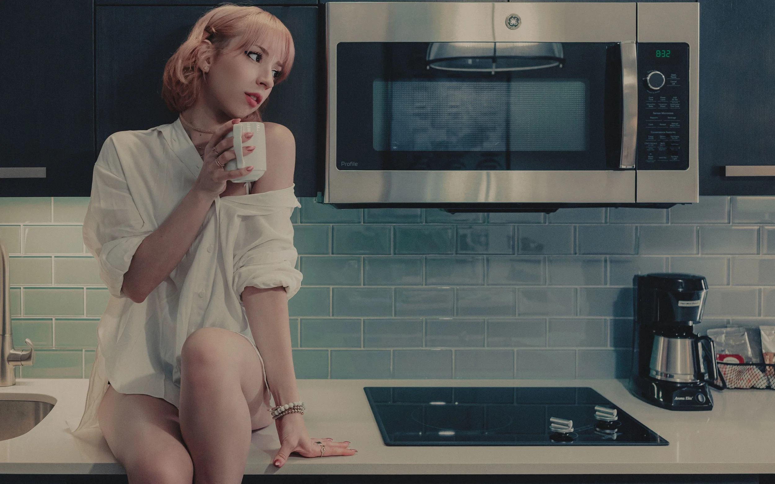 A woman with pink hair sitting on a kitchen counter, holding a white mug, wearing an open shirt and lingerie, with a microwave and kitchen appliances visible in the background.