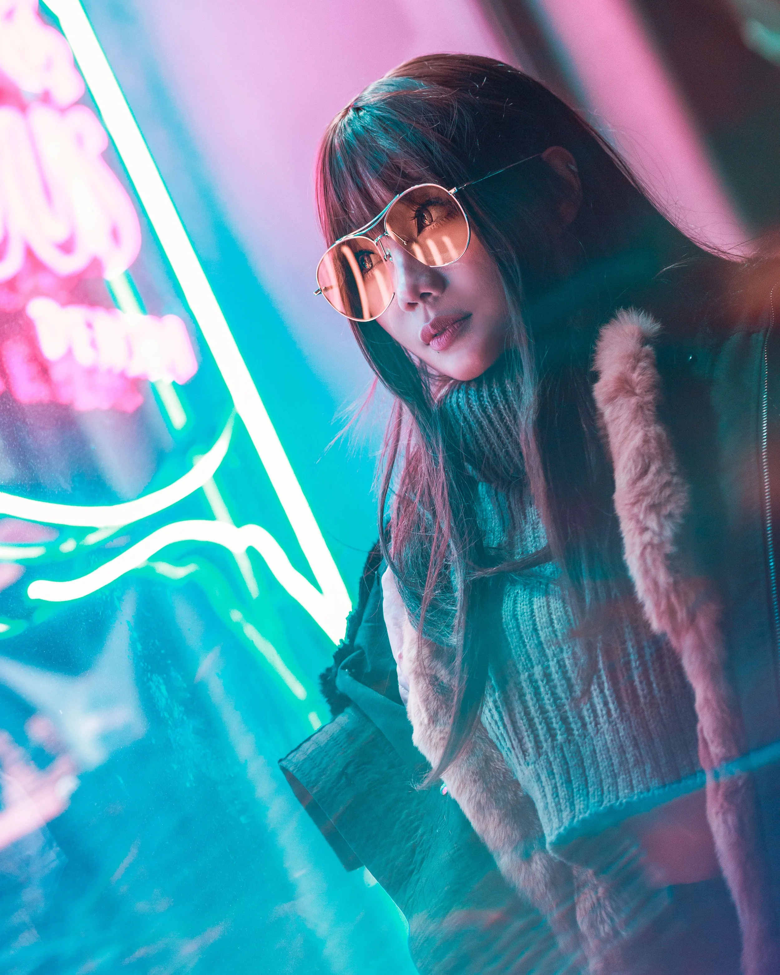 A woman with long dark hair and sunglasses posing in front of colorful neon signs at night.