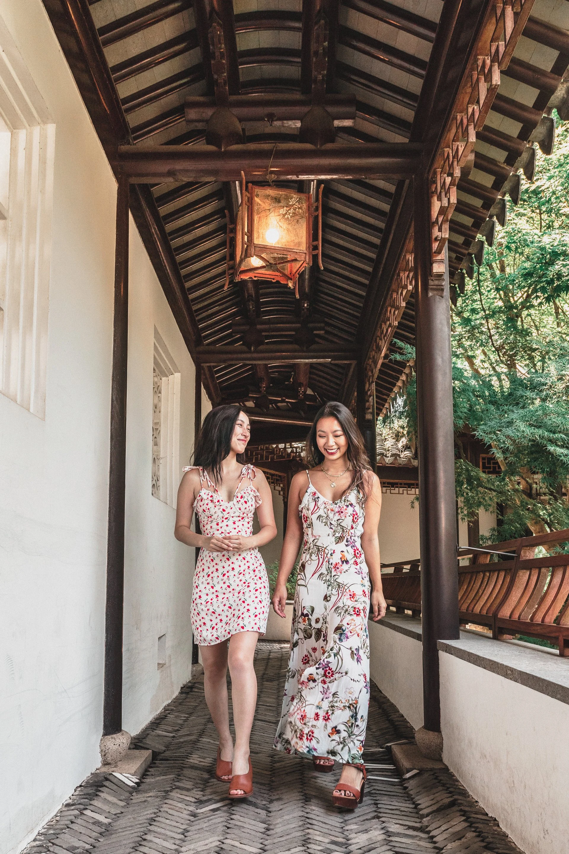 Two women wearing floral dresses walk along a paved pathway underneath a wooden covered corridor, smiling and talking to each other, with greenery visible in the background.