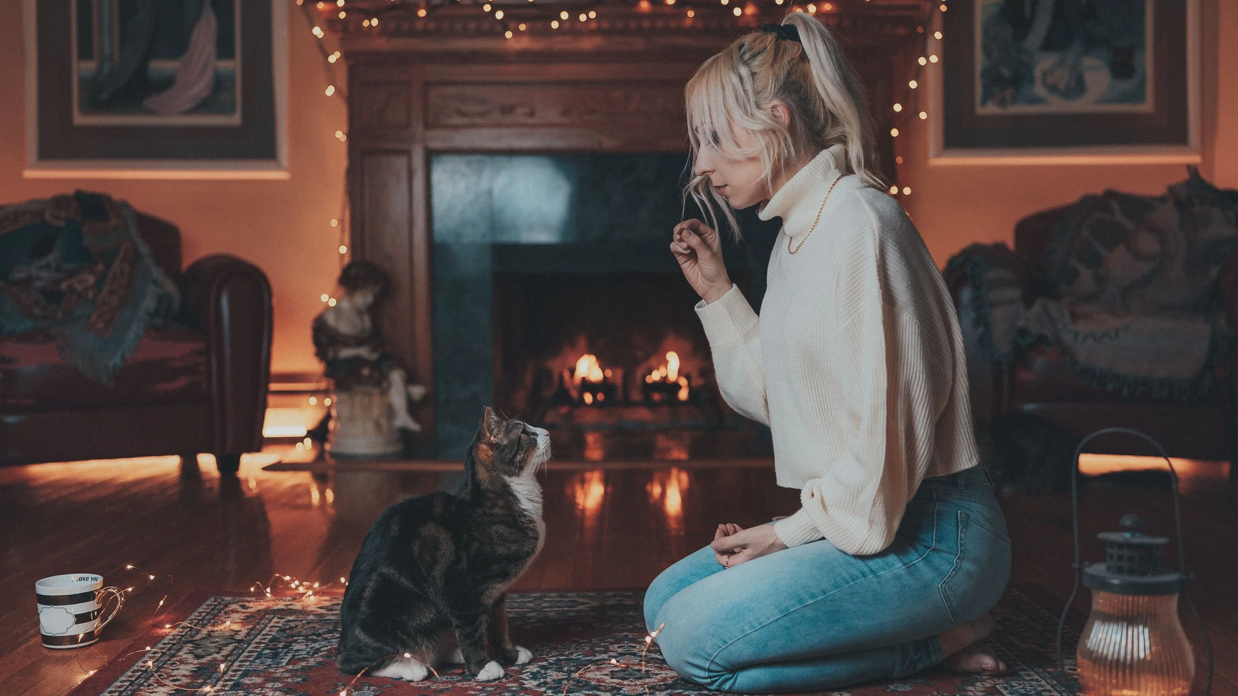 A woman kneeling on a rug in front of a fireplace, interacting with a tabby cat. The room is decorated with fairy lights, and there are paintings on the wall, a mug on the floor, and a basket with blankets.